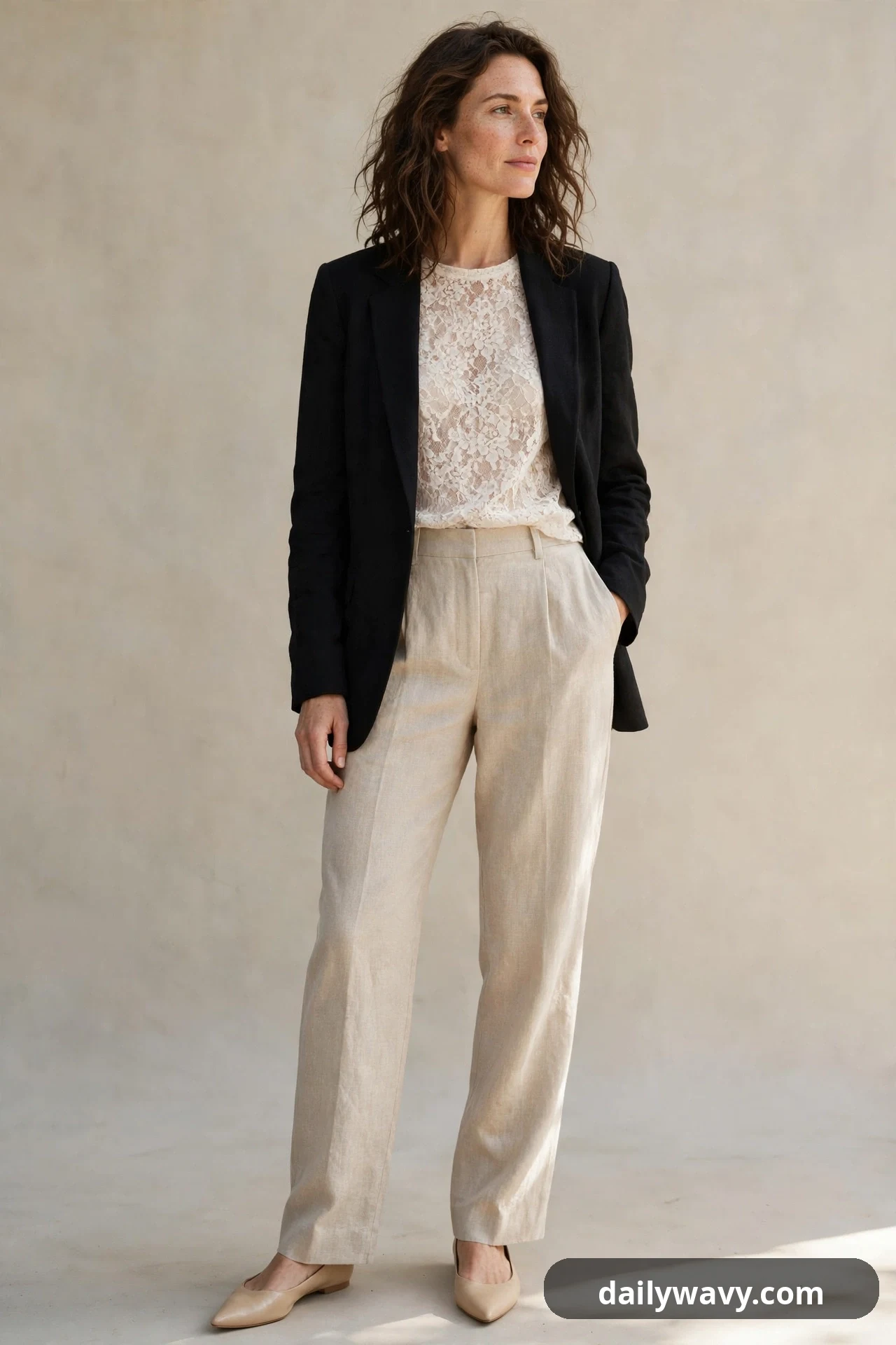 A woman wears an ivory opaque lace top under a tailored black blazer, with sand linen trousers and nude pointed flats