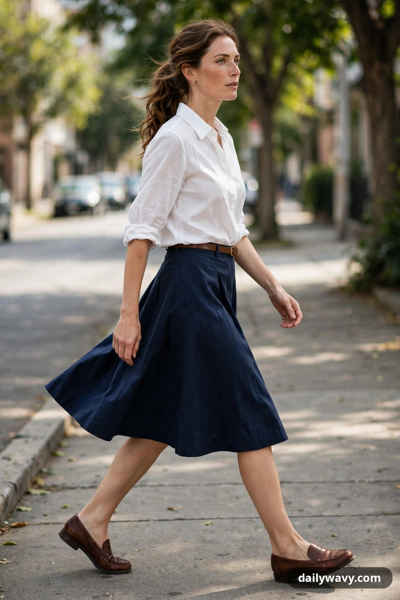 A woman walks in a navy A-line skirt and a white collared blouse tucked in with a thin brown belt, wearing dark brown loafers.