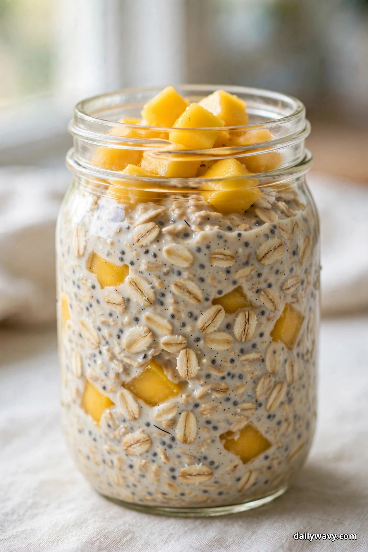 A glass jar of mango overnight oats speckled with chia seeds is topped with a mound of golden diced mango