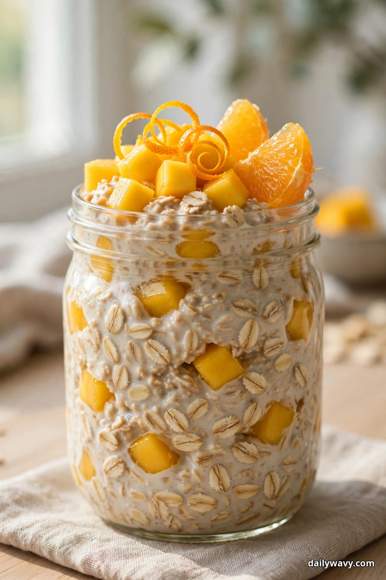 A glass jar of mango overnight oats is topped with golden diced mango, curled strips of orange zest and orange segments