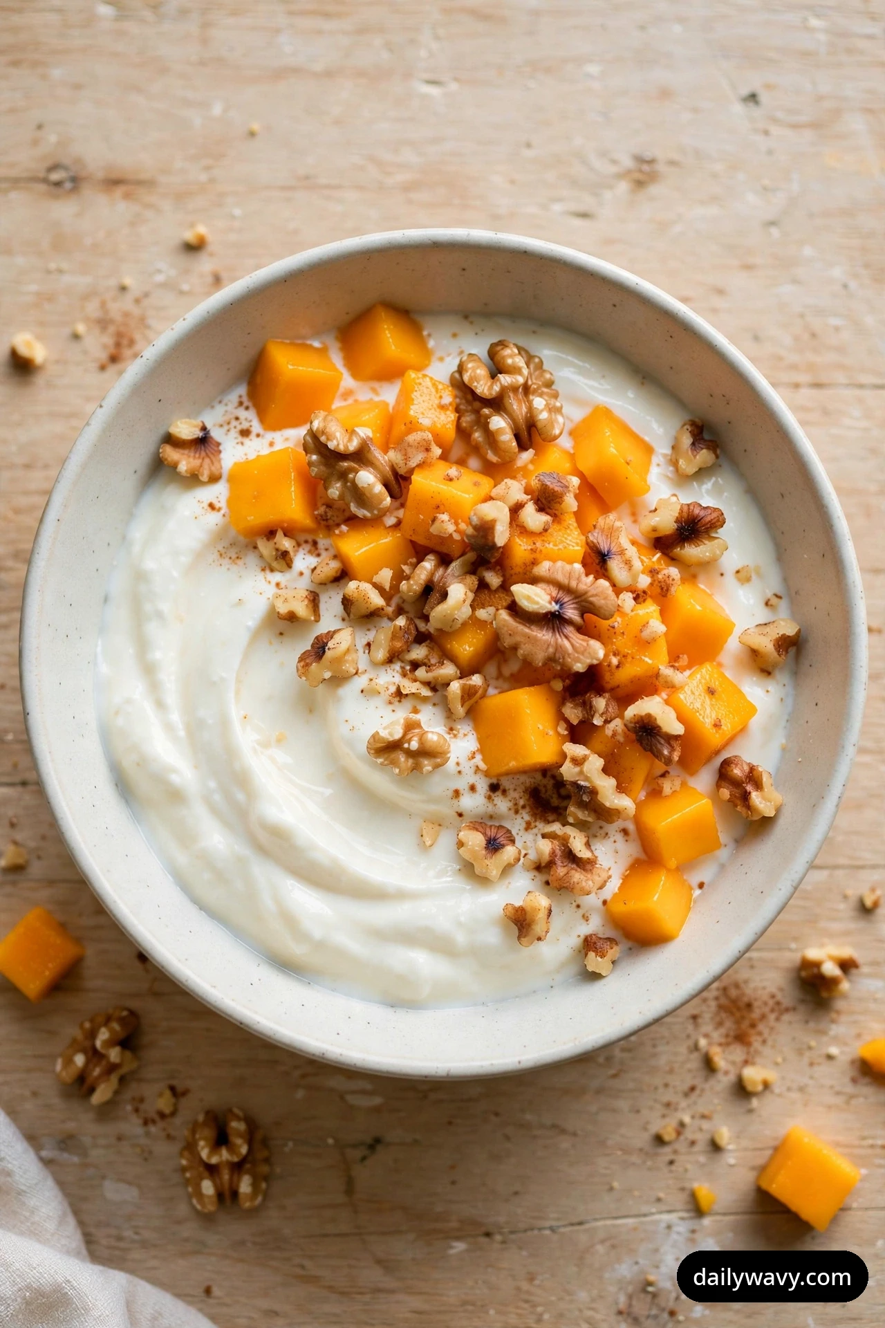 An overhead view shows a ceramic bowl of swirled yogurt topped with mango cubes, chopped walnuts, and a dusting of cinnamon.
