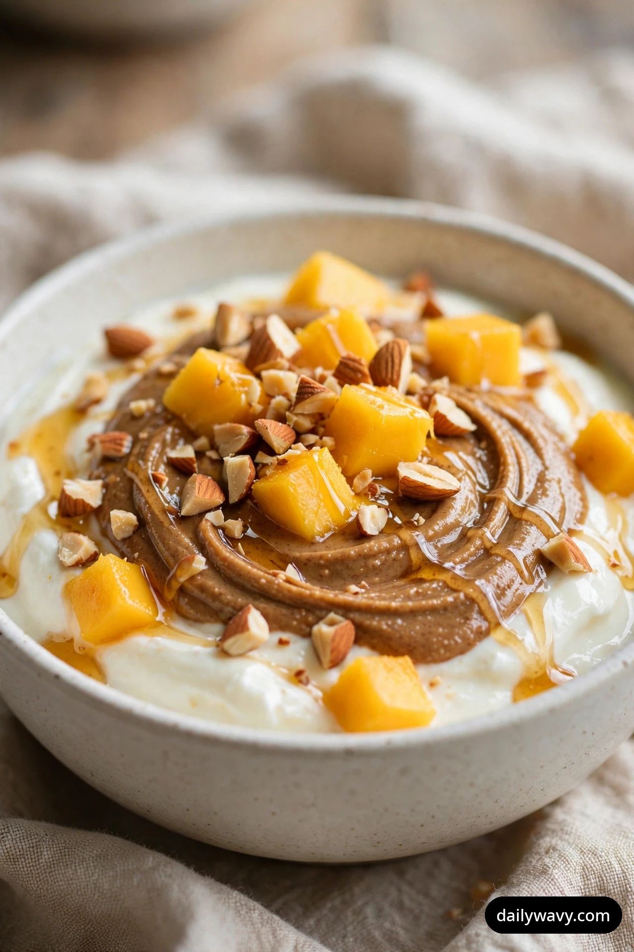 A close-up shows a bowl of yogurt with a thick swirl of almond butter, topped with mango cubes, chopped almonds, and a honey drizzle.