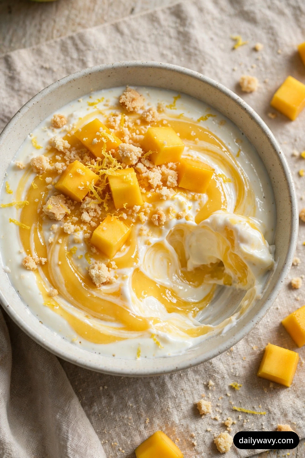 An overhead view shows a bowl of yogurt marbled with lemon curd swirls, topped with mango cubes, shortbread crumbs, and fine lemon zest.