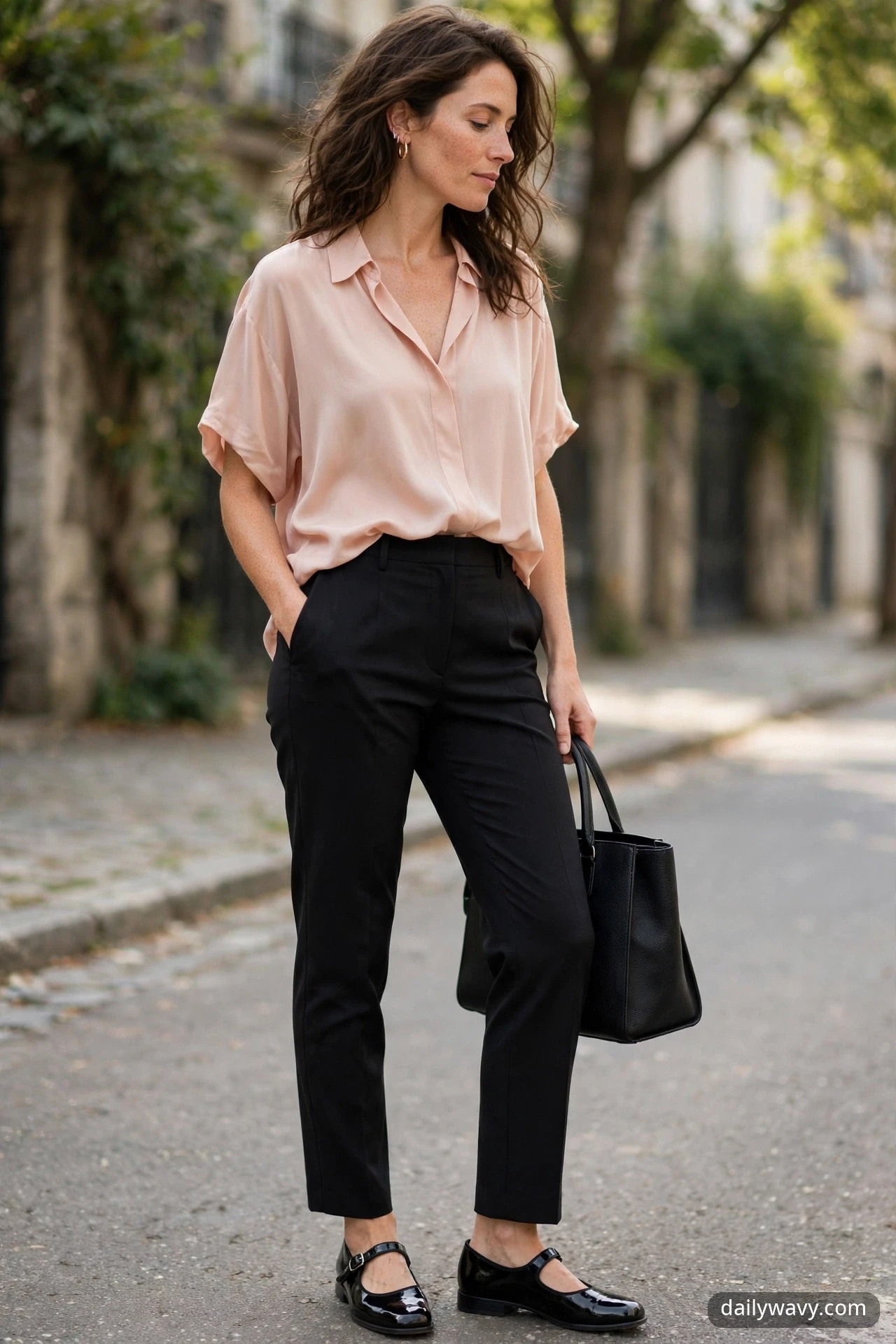 Adult model in a relaxed pose wearing black tailored trousers, a blush soft blouse and black patent Mary Jane shoes.