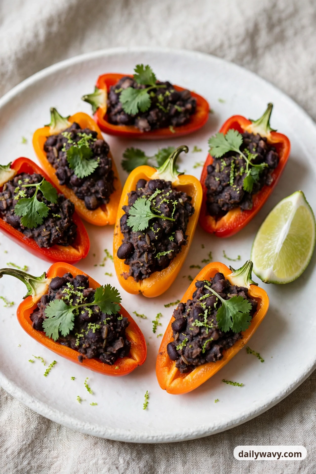 Mini sweet pepper halves on a white plate are filled with mashed black beans and topped with cilantro leaves and lime zest, with a lime wedge beside them on the plate.