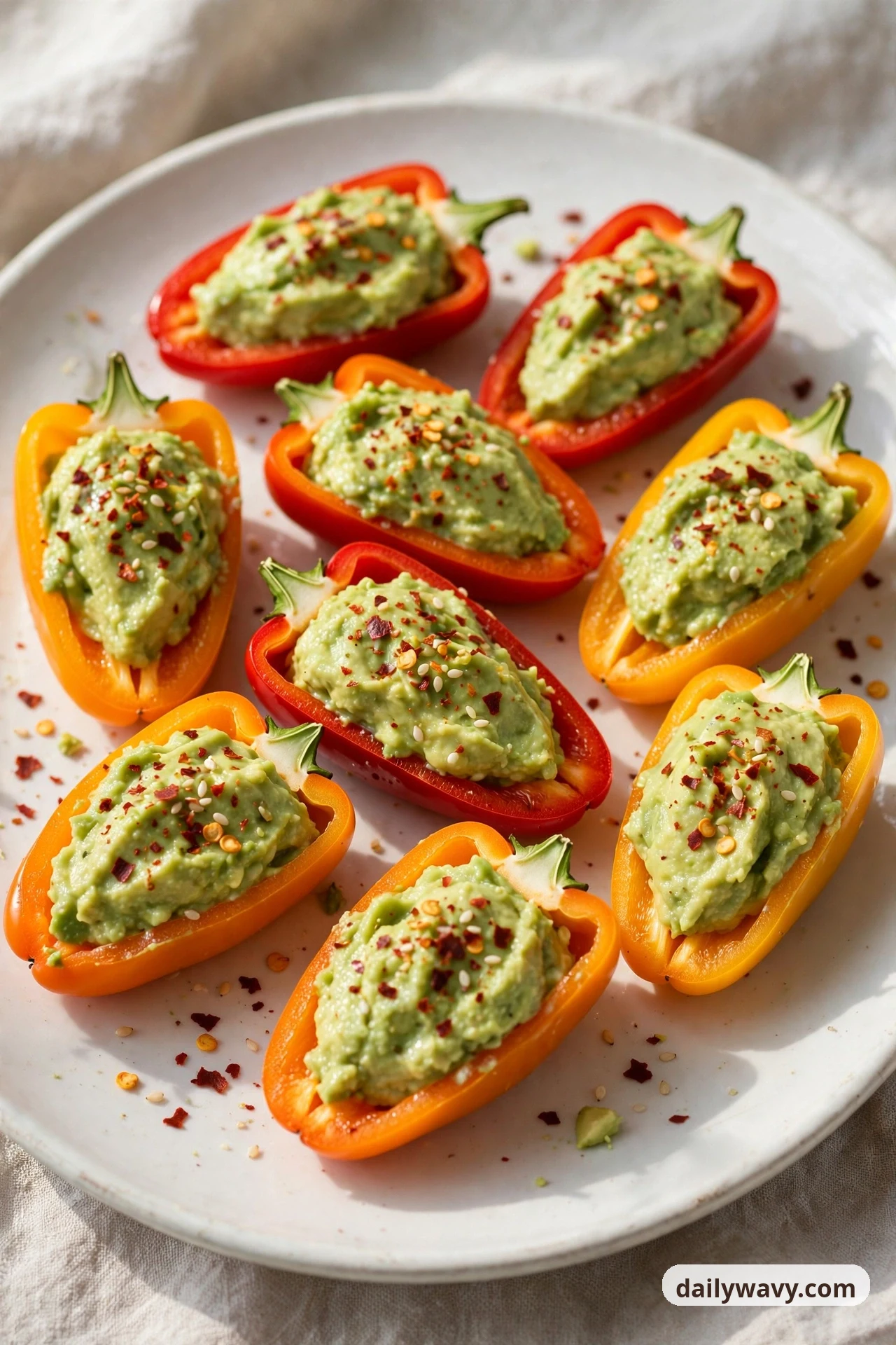 Mini sweet pepper halves on a white plate are filled with mashed avocado and sprinkled with chili flakes and sesame seeds.