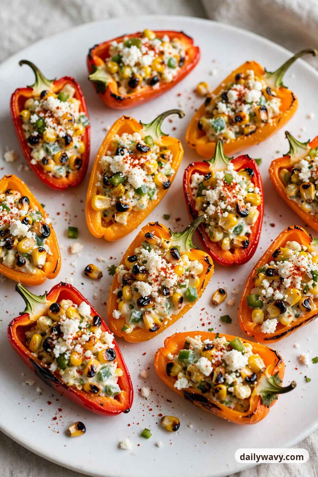 Mini sweet pepper halves on a white plate are filled with a charred corn and jalapeno mixture and topped with crumbled cotija and a dusting of chili powder.