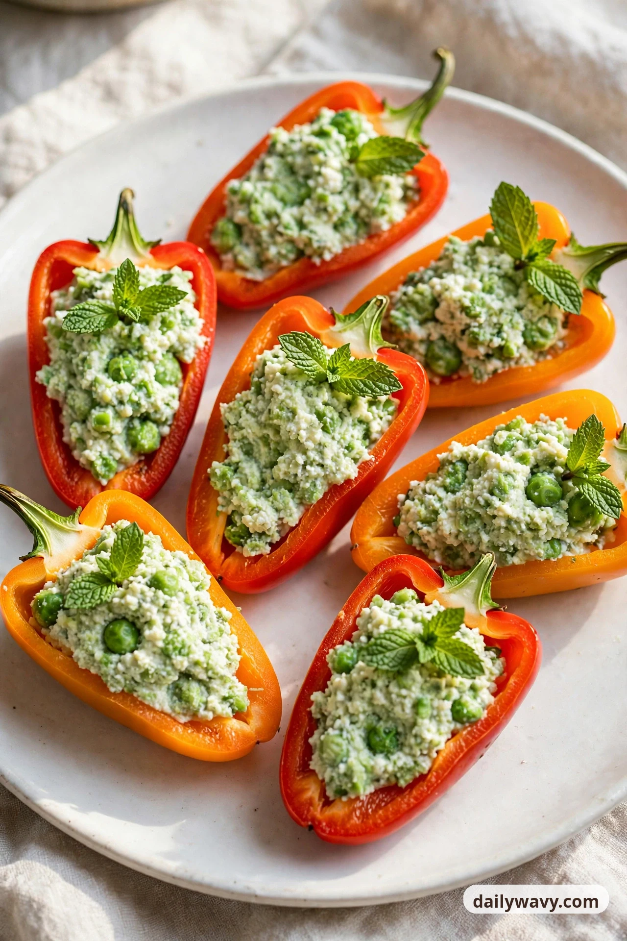Mini sweet pepper halves on a white plate are filled with a crushed pea and ricotta mixture with whole peas visible, each topped with fresh mint leaves.