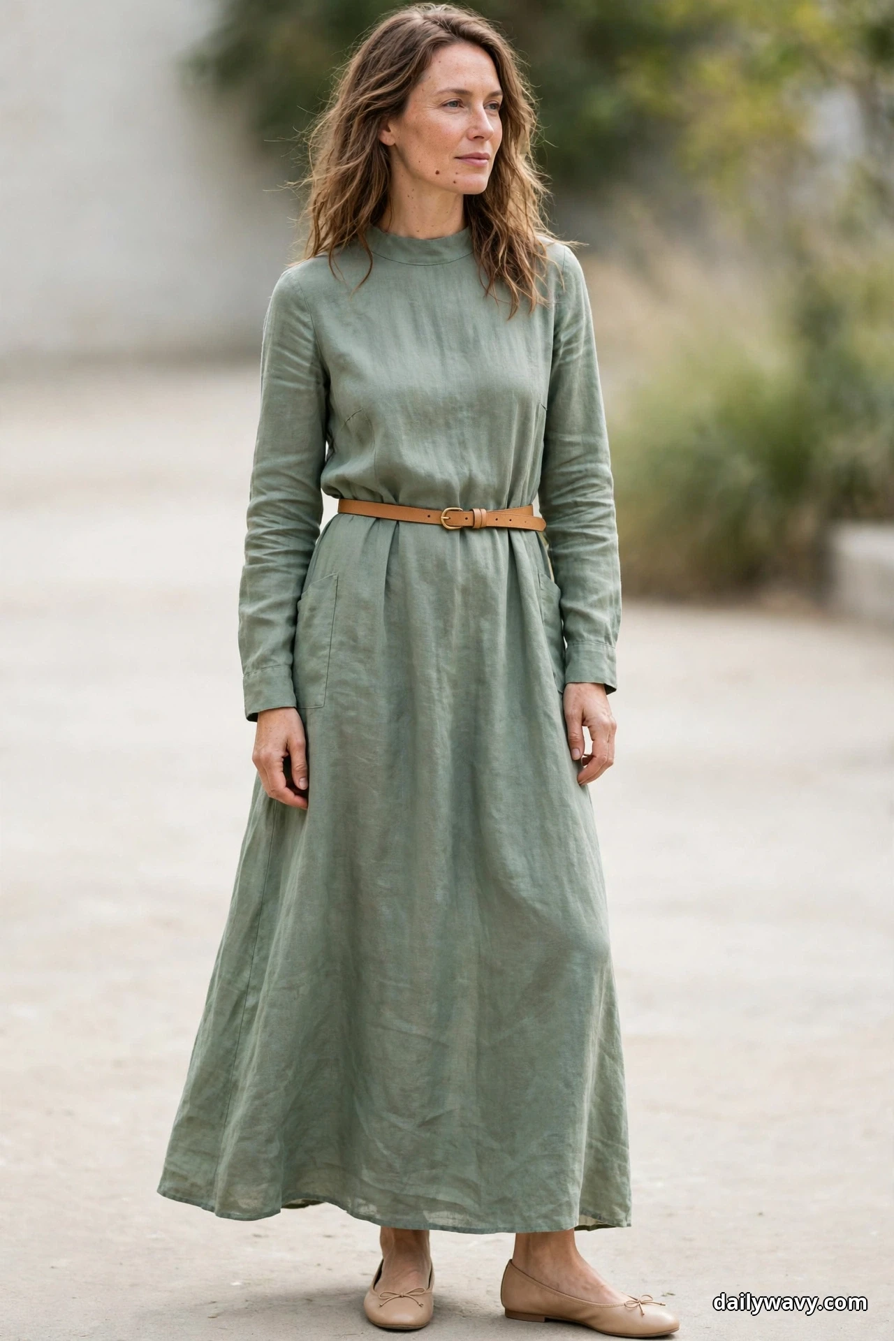 A woman stands outdoors wearing a sage-green long-sleeve maxi dress with a high round neckline, cinched with a narrow tan leather belt, and nude ballet flats