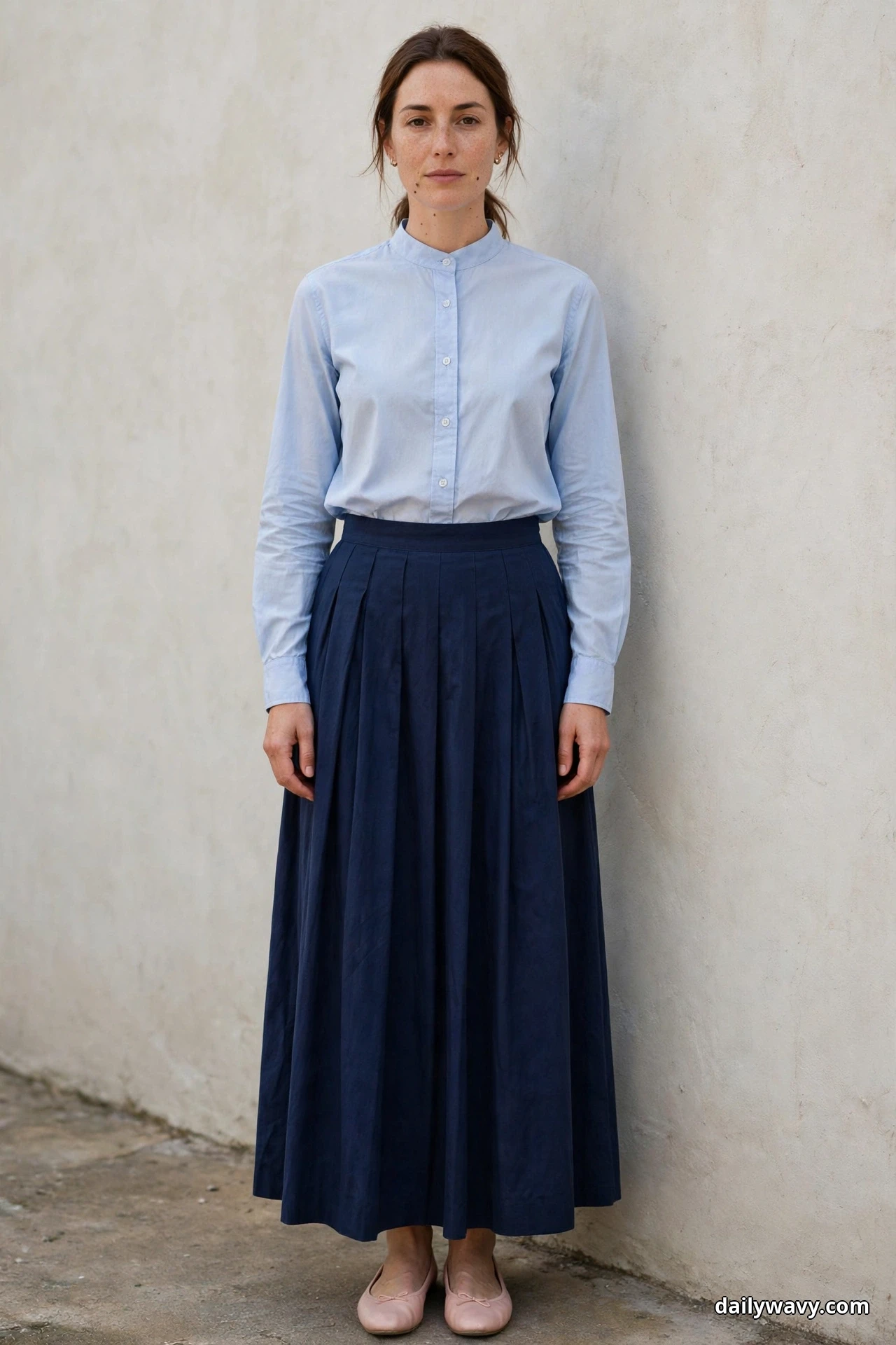A woman stands against a textured wall wearing a light-blue long-sleeve button-up shirt tucked into a navy pleated floor-length skirt, with blush ballet flats