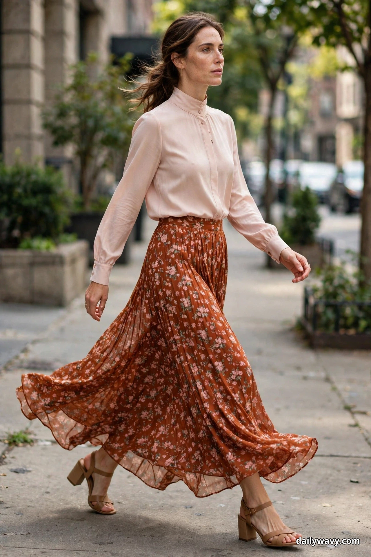 A woman walks along a city sidewalk wearing a blush high-neck long-sleeve blouse tucked into a flowing rust floral-print maxi skirt, with tan block-heel sandals