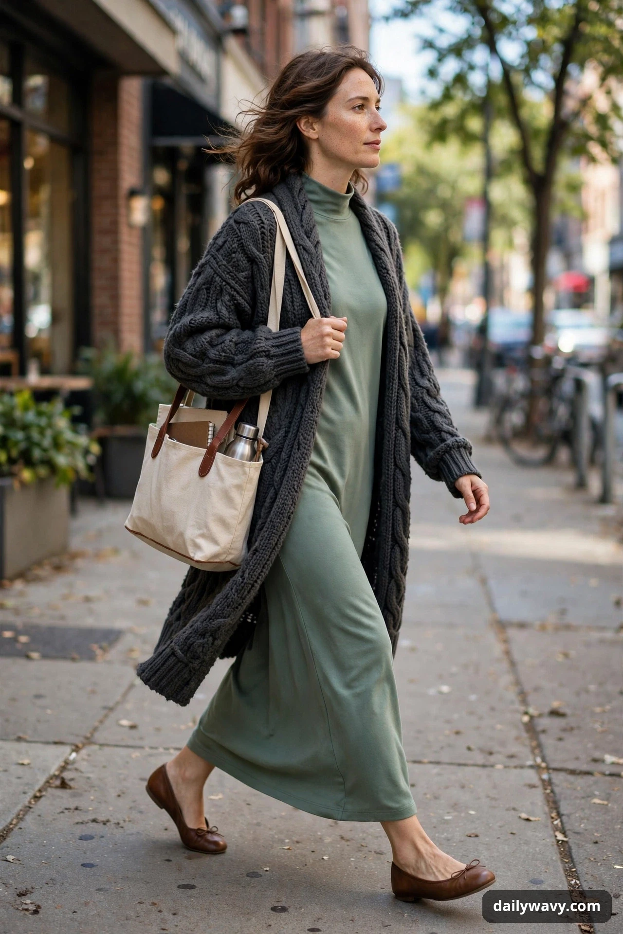 A woman walks along a city sidewalk in a sage green full-length jersey dress under a charcoal chunky cable-knit cardigan, wearing brown ballet flats and carrying a canvas tote.