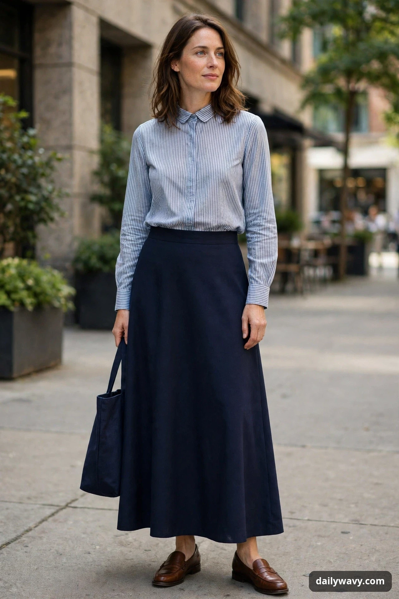 A woman stands outdoors wearing a blue-and-white striped collared blouse tucked into a navy A-line maxi skirt, with brown loafers and a navy tote in her hand.