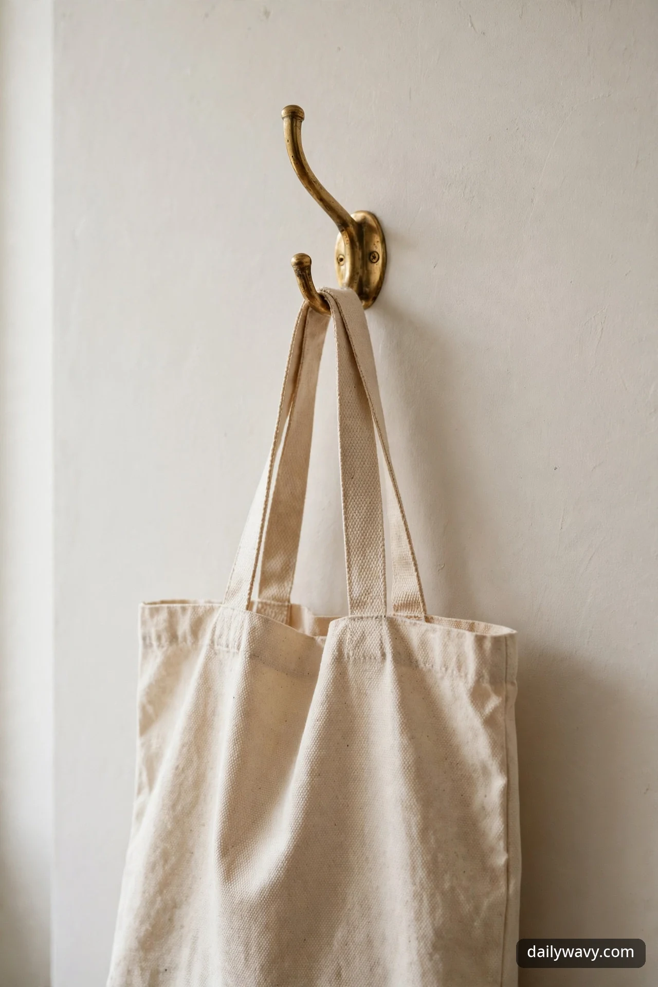 A two-arm brass wall hook holds a natural canvas tote bag against a warm off-white hallway wall.