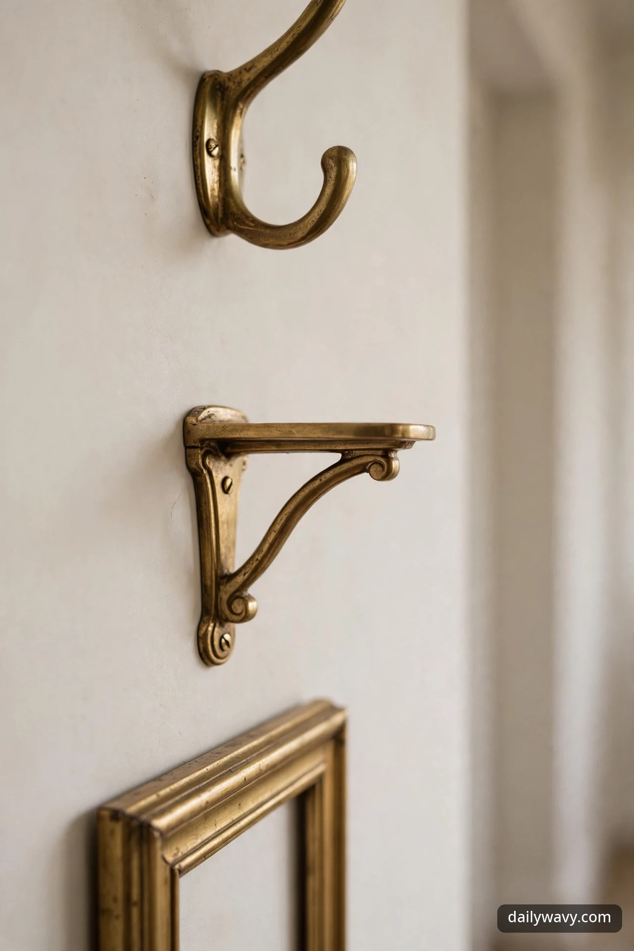 A close-up shows an aged-brass wall hook, a matching ornate brass shelf bracket, and the corner of a brass picture frame sharing one finish on a cream wall.
