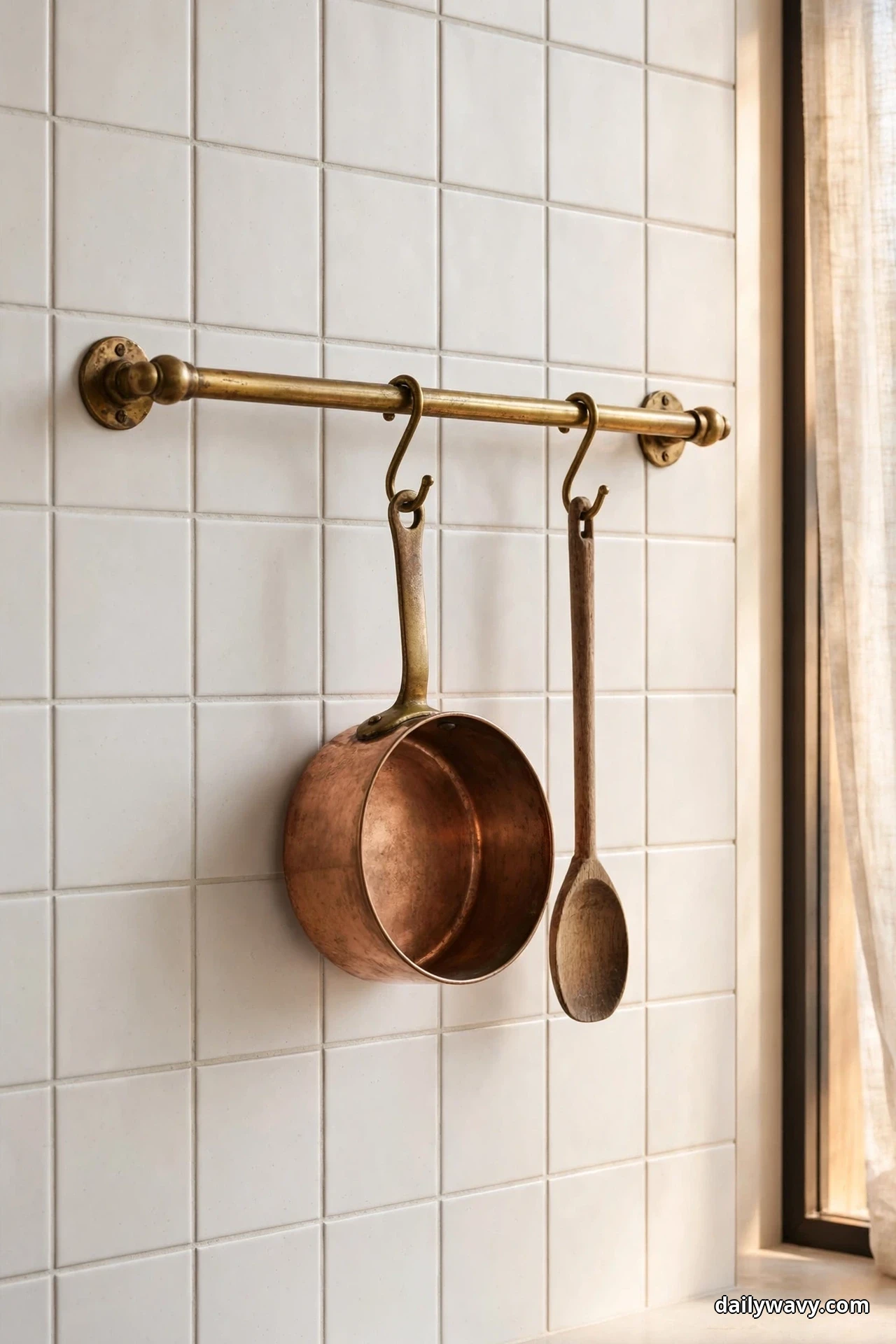 An antique brass pot rail mounted across a white square tile backsplash holds a copper saucepan and a wooden spoon on hooks