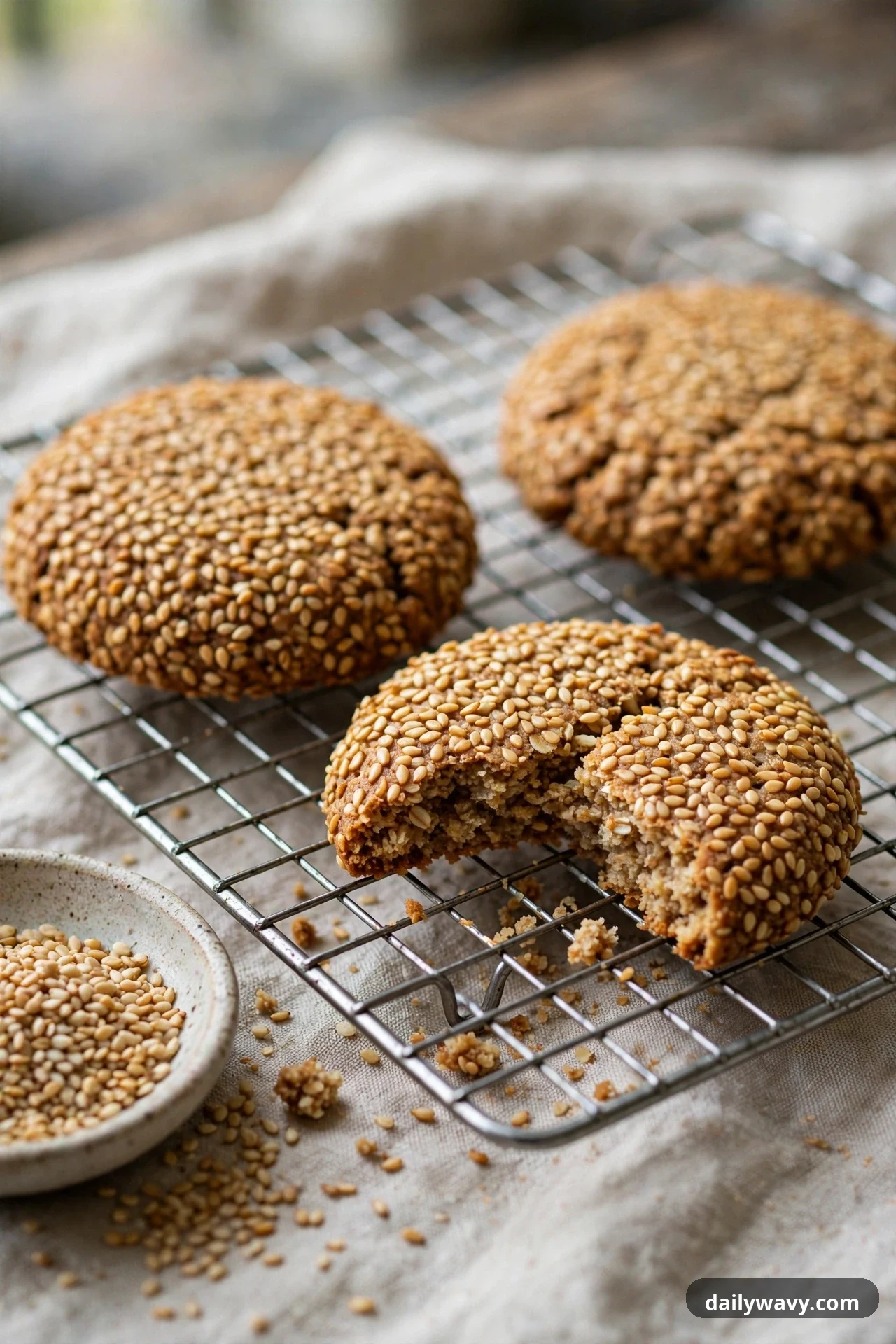 Oatmeal cookies fully coated in toasted sesame seeds rest on a wire cooling rack over a linen cloth, with one cookie broken open and a small bowl of sesame seeds beside the rack.