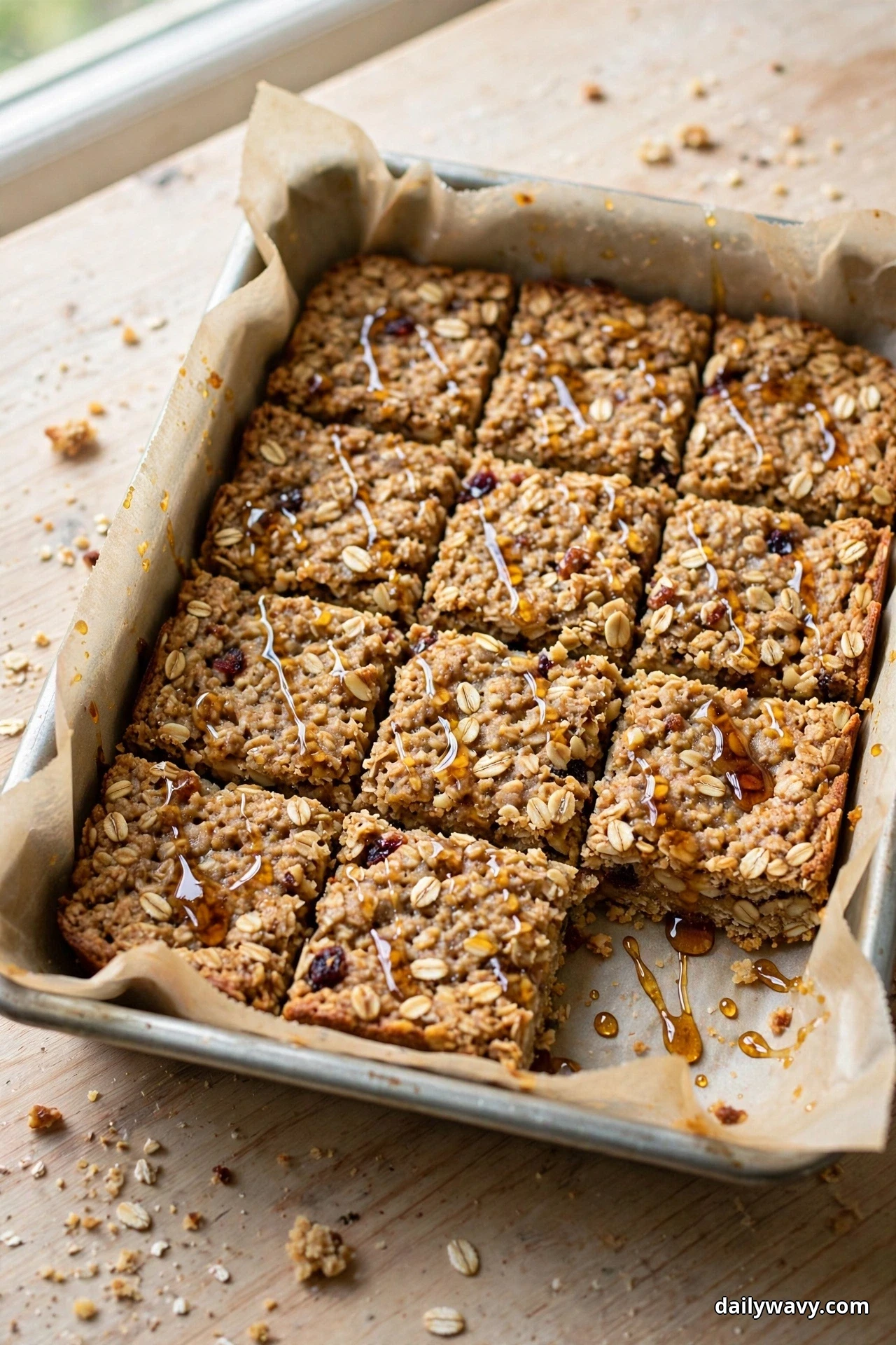 Cut oatmeal bars with a thin glossy maple glaze streaked across their tops sit in a parchment-lined baking pan on a wooden table