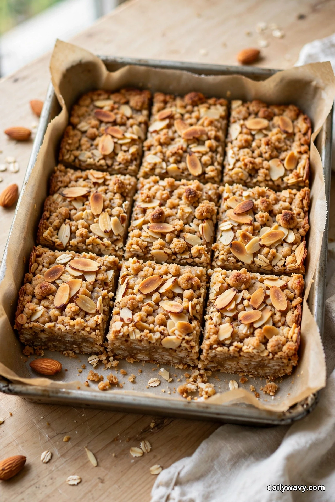 Cut oatmeal bars topped with toasted sliced almonds sit in a parchment-lined baking pan, with whole almonds scattered on the wooden table beside the pan