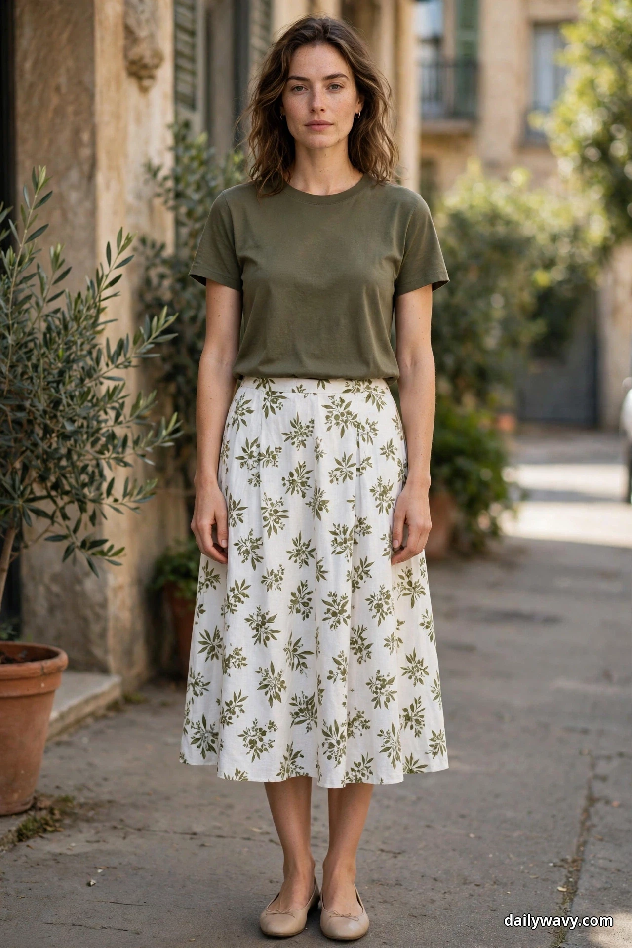 A woman wearing an olive crew-neck tee and a white midi skirt with an olive floral print stands full length on a village street in nude ballet flats