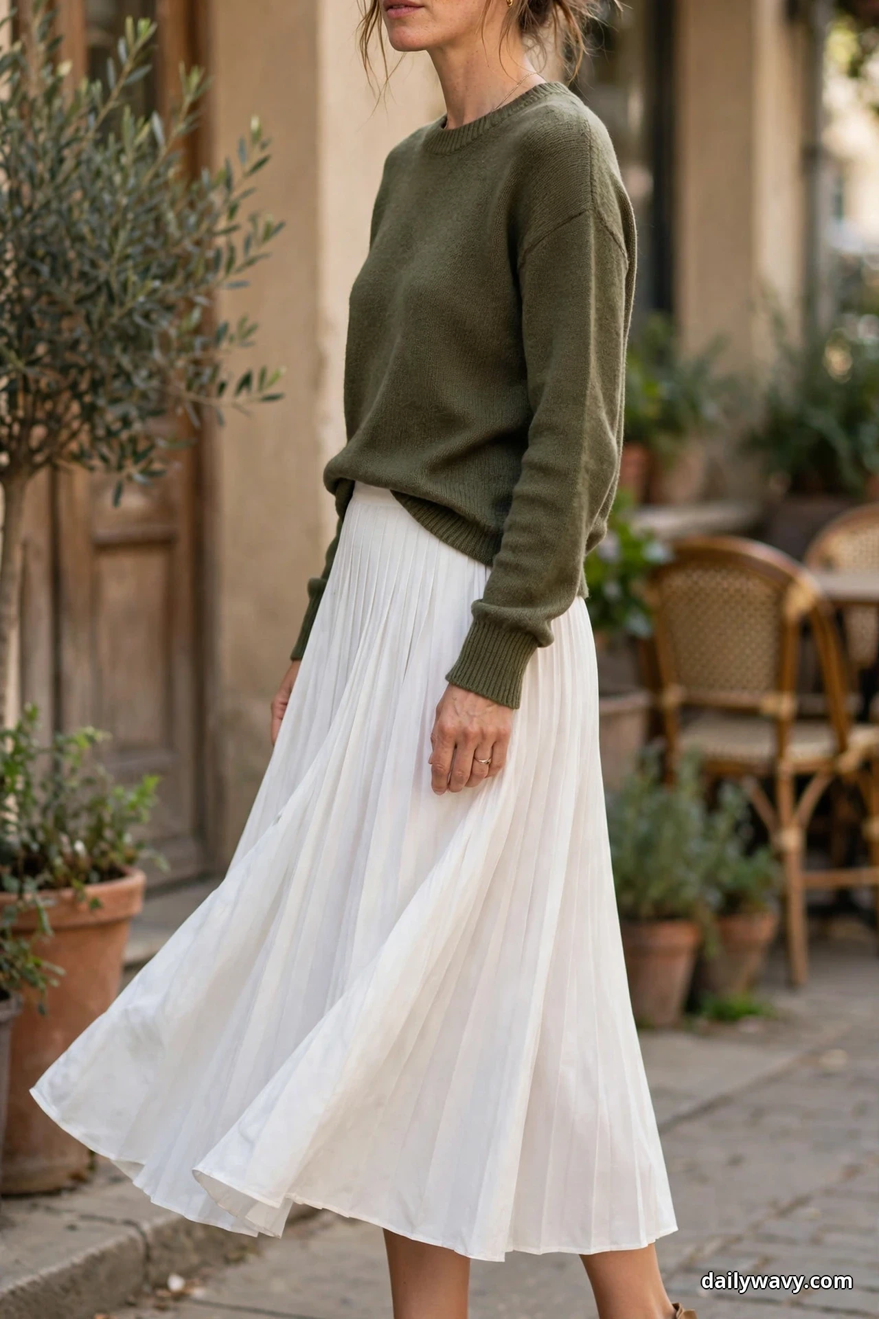 A woman wearing an olive crewneck sweater and a white pleated midi skirt is shown from the chin down at an outdoor cafe with the frame ending below the skirt hem and her footwear out of view
