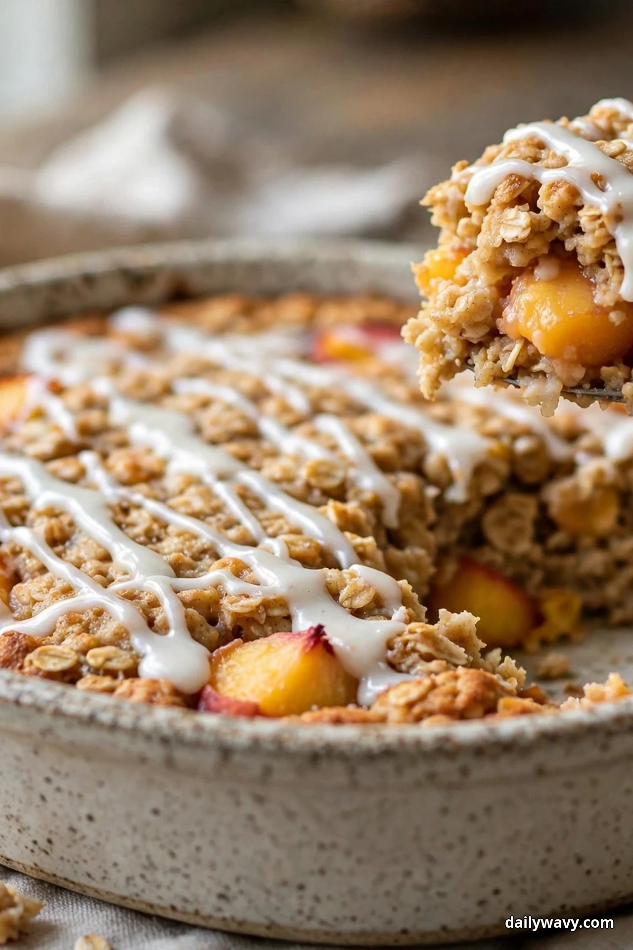 A round ceramic dish of peach baked oatmeal is drizzled with white vanilla glaze in zigzags, with a spoonful lifted at the edge showing the peach chunks inside