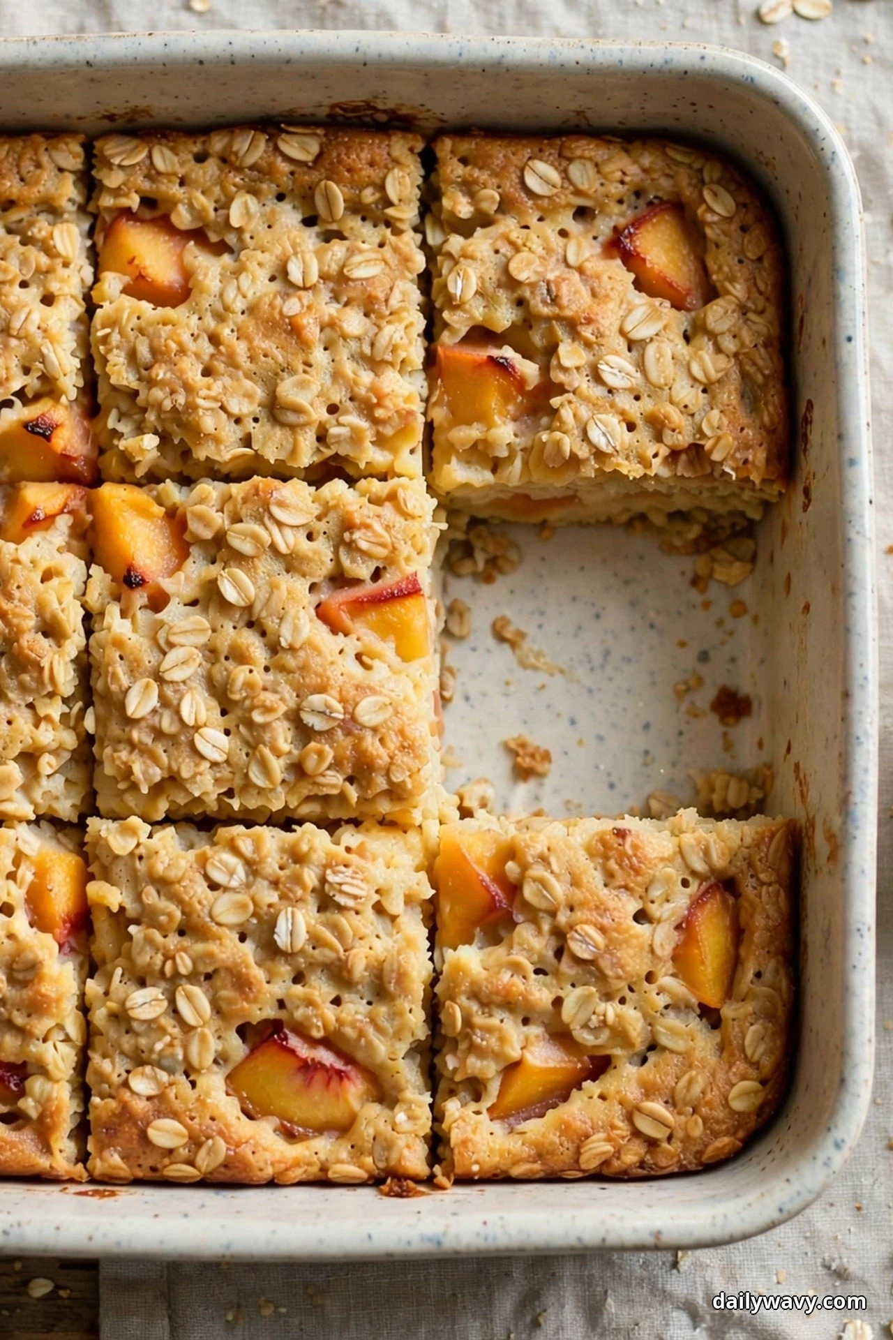 Seen from above, a square ceramic dish of peach baked oatmeal is cut into firm squares studded with peach chunks, with one square lifted out