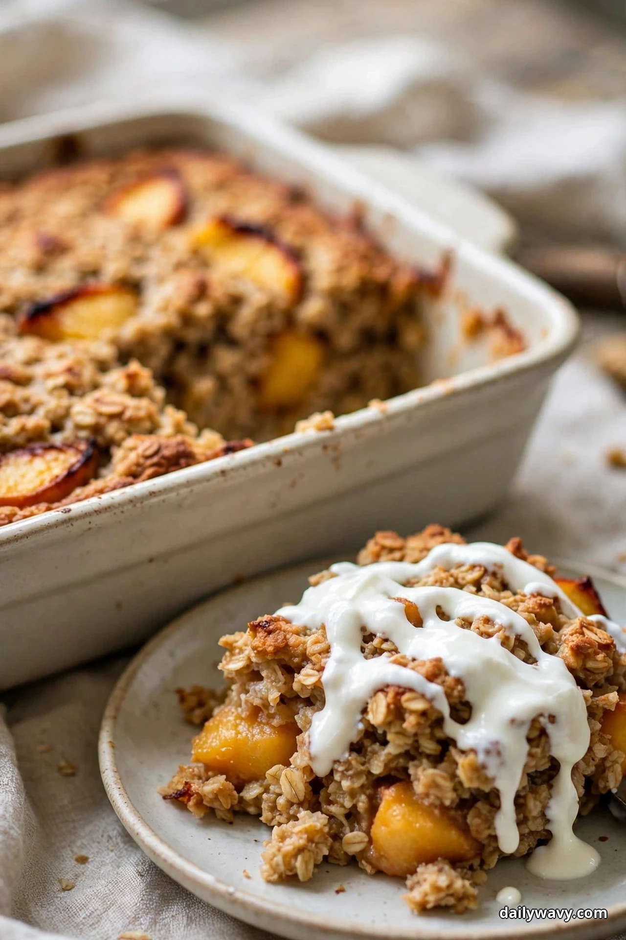 A slice of peach baked oatmeal on a plate is spooned over with a white yogurt drizzle, with the baking dish of peach oatmeal behind it