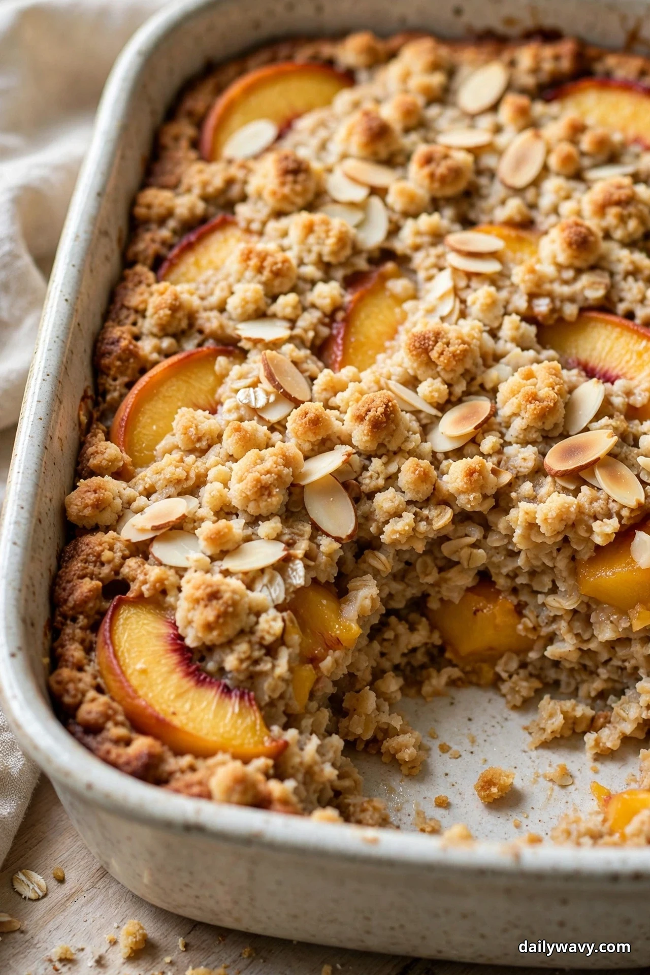 A ceramic baking dish of peach baked oatmeal is covered with golden crumble clusters and sliced almonds over peach slices, with one serving lifted out