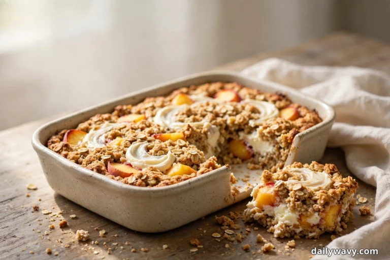 A ceramic baking dish of peach baked oatmeal with cream-cheese swirls ribboned across the golden top sits on a wooden table beside a linen cloth, with one slice lifted out showing the peach and cream-cheese layers