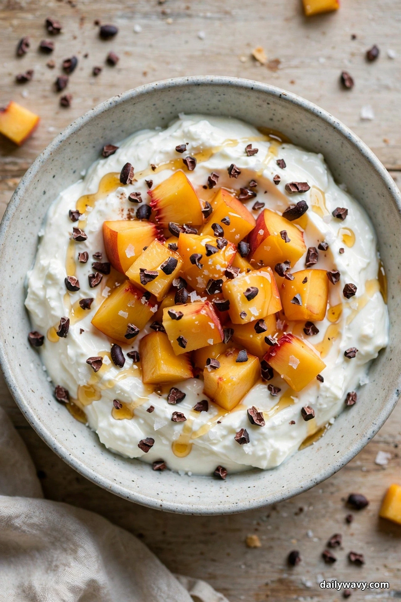 Seen from directly above, a bowl of yogurt piled with diced peaches and scattered cocoa nibs, flaky salt and a honey drizzle sits on a wooden table with more nibs and peach pieces around it