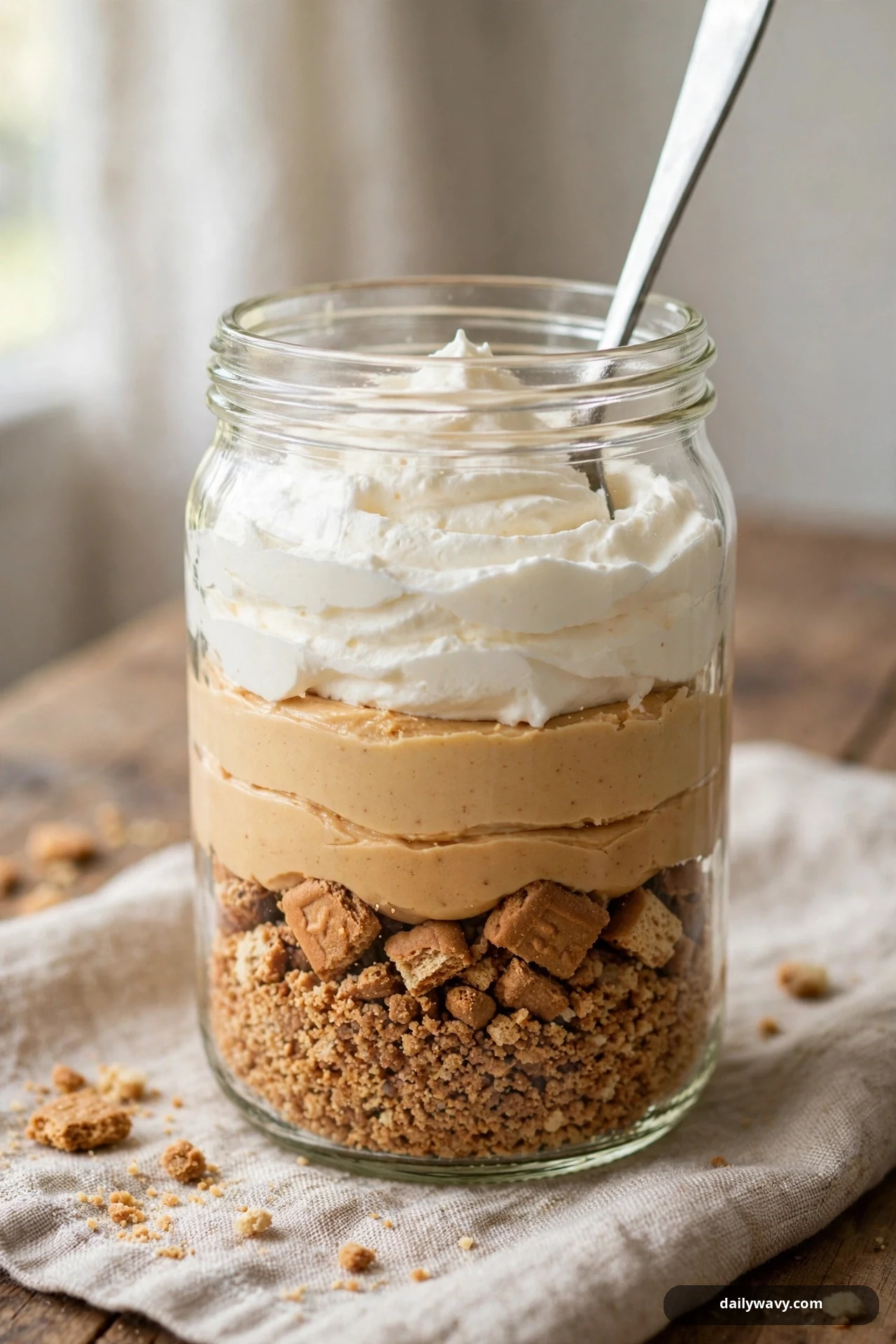 A glass jar shows stripes of biscuit crumbs and chunks, peanut butter cream, and whipped cream, with a long spoon standing in the jar.