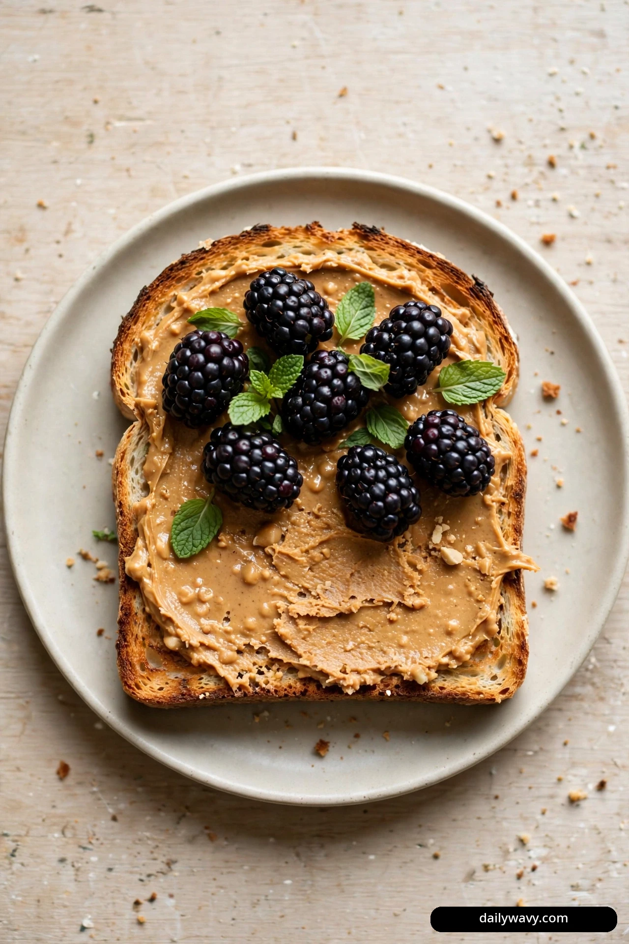 A top-down view shows a slice of sourdough toast spread with peanut butter and topped with whole blackberries and fresh mint leaves.