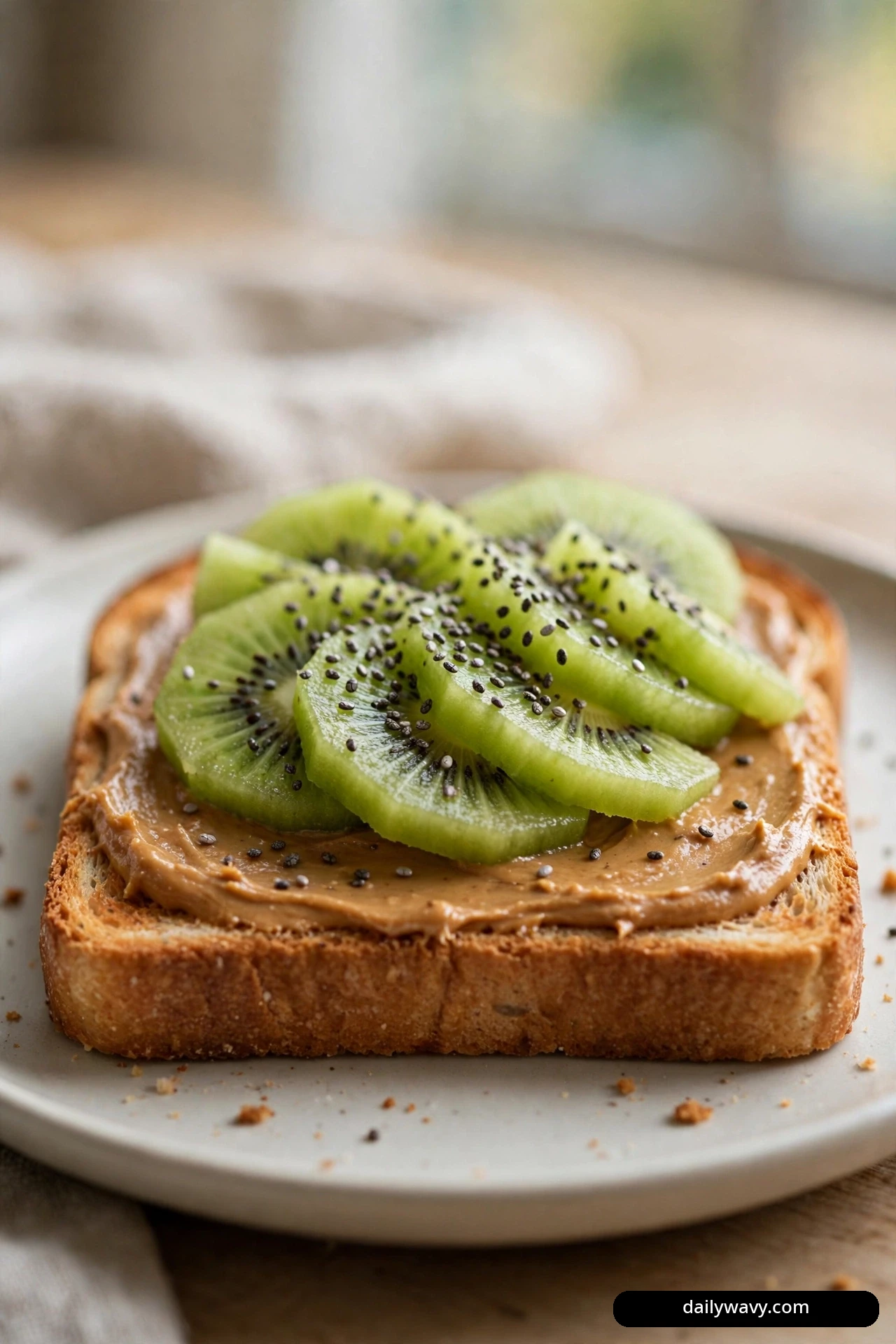 A close view shows a slice of whole wheat toast spread with peanut butter and topped with fanned kiwi slices and a sprinkle of chia seeds.