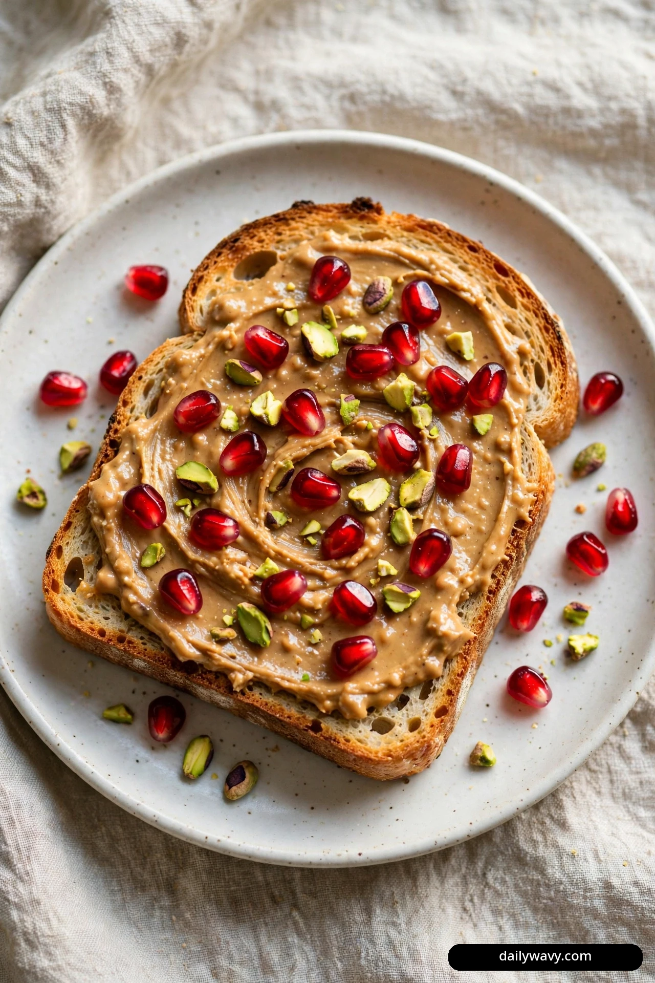 A top-down view shows a slice of sourdough toast spread with peanut butter and scattered with pomegranate arils and chopped pistachios.