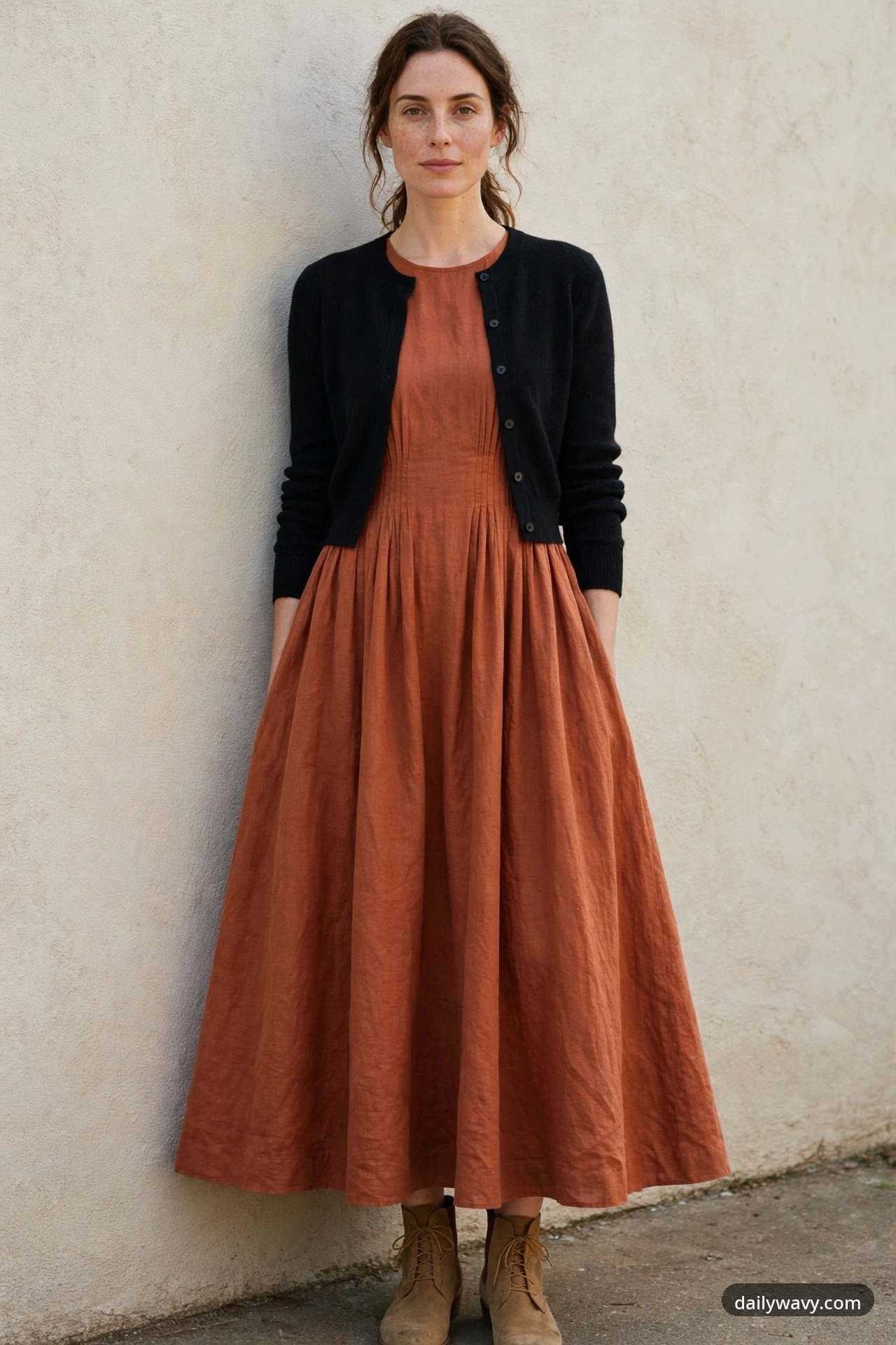 A petite woman stands against a stucco wall wearing a terracotta A-line maxi dress with a black cropped buttoned cardigan and tan lace-up ankle boots.
