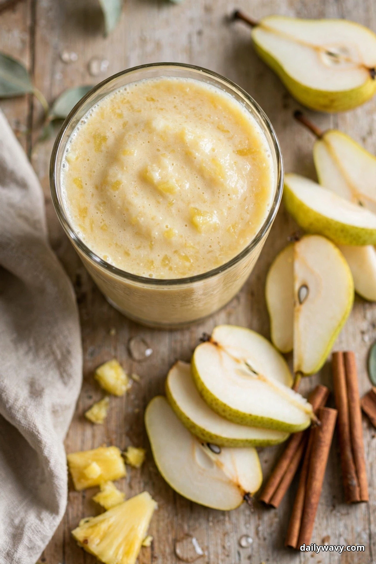 Seen from above, a glass of pale creamy pineapple pear smoothie sits on a wooden table surrounded by pear slices, a halved pear, cinnamon sticks and pineapple chunks