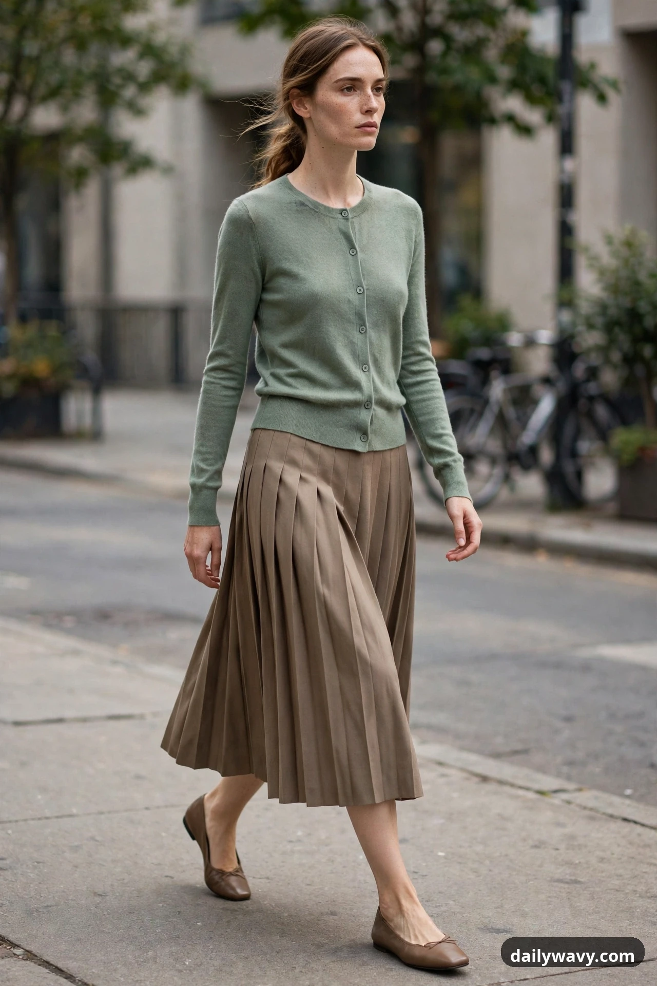 A model walks along a city sidewalk in a sage green buttoned cardigan worn as a top with a taupe pleated midi skirt and taupe ballet flats.