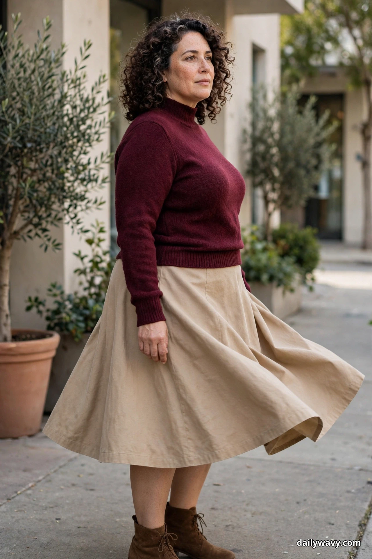 A woman turns on a sidewalk wearing a burgundy long-sleeve mock-neck knit top with a beige A-line midi skirt that flares as she moves, with brown suede lace-up ankle boots