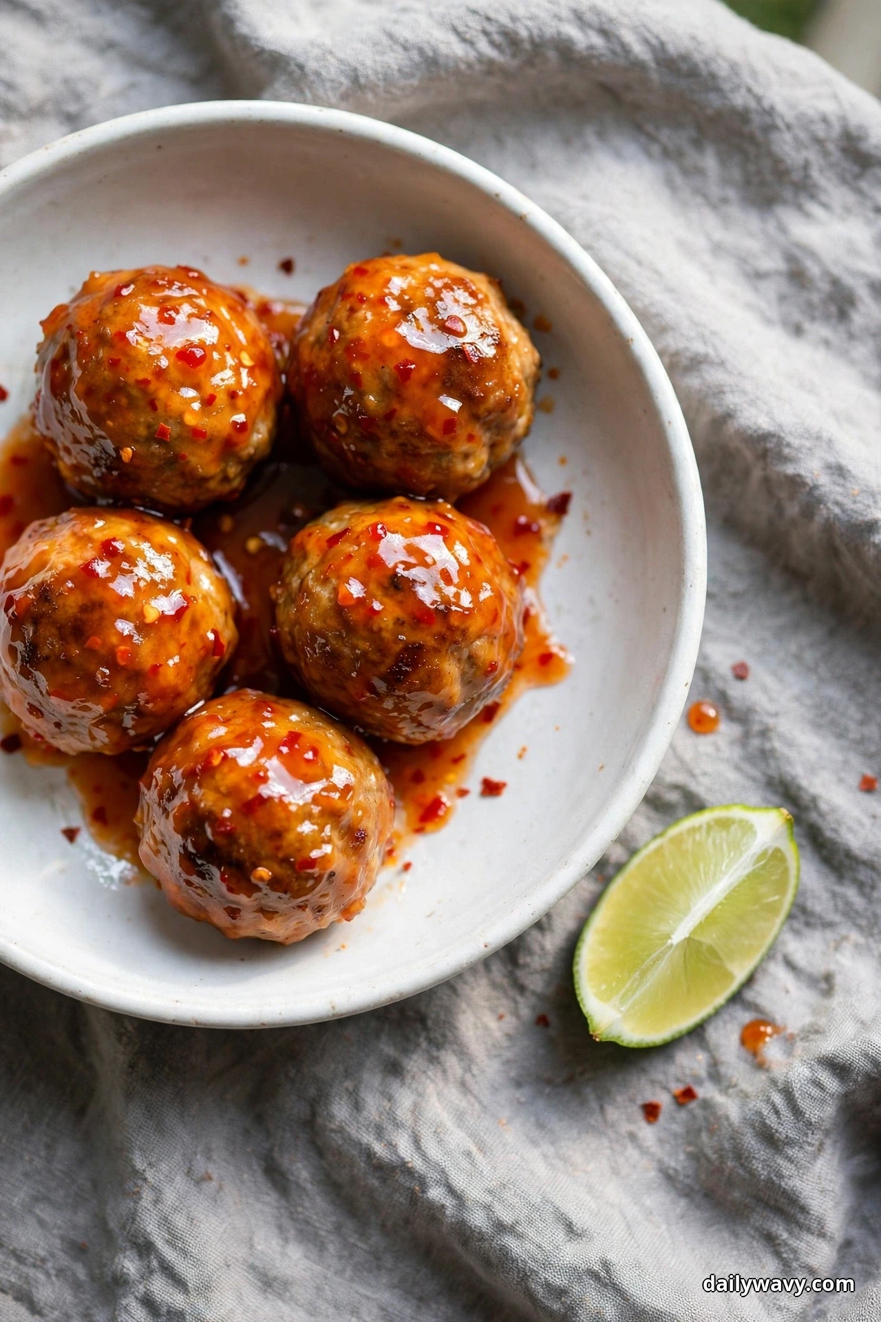 Seen from above, a white bowl holds pork meatballs coated in a glossy orange-red sweet chili glaze flecked with chili, resting on a gray linen cloth
