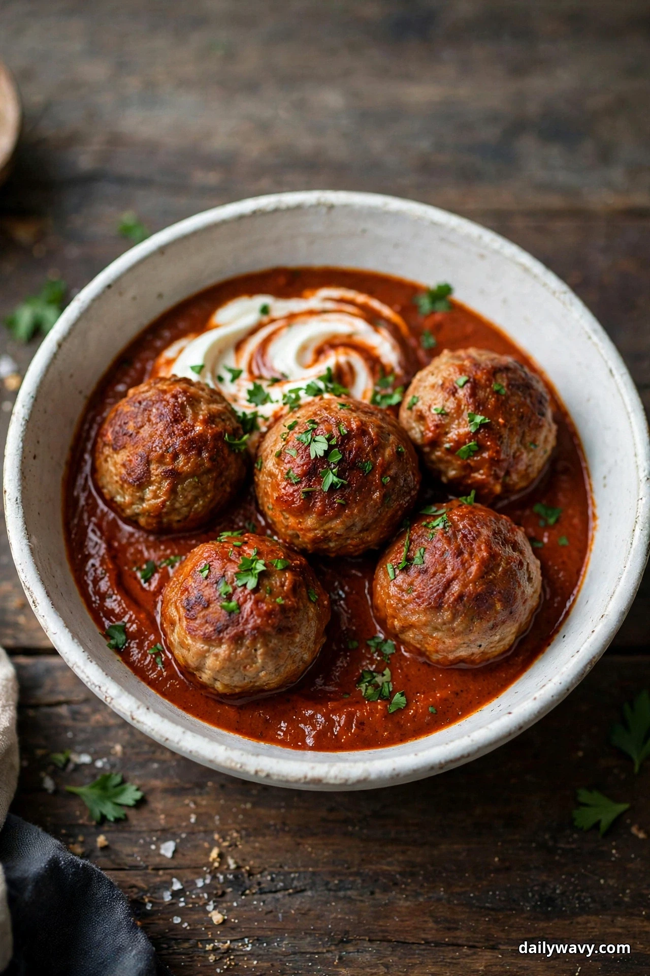 A deep bowl holds pork meatballs in a dark red paprika sauce with a swirl of sour cream on the surface, on a dark wooden table