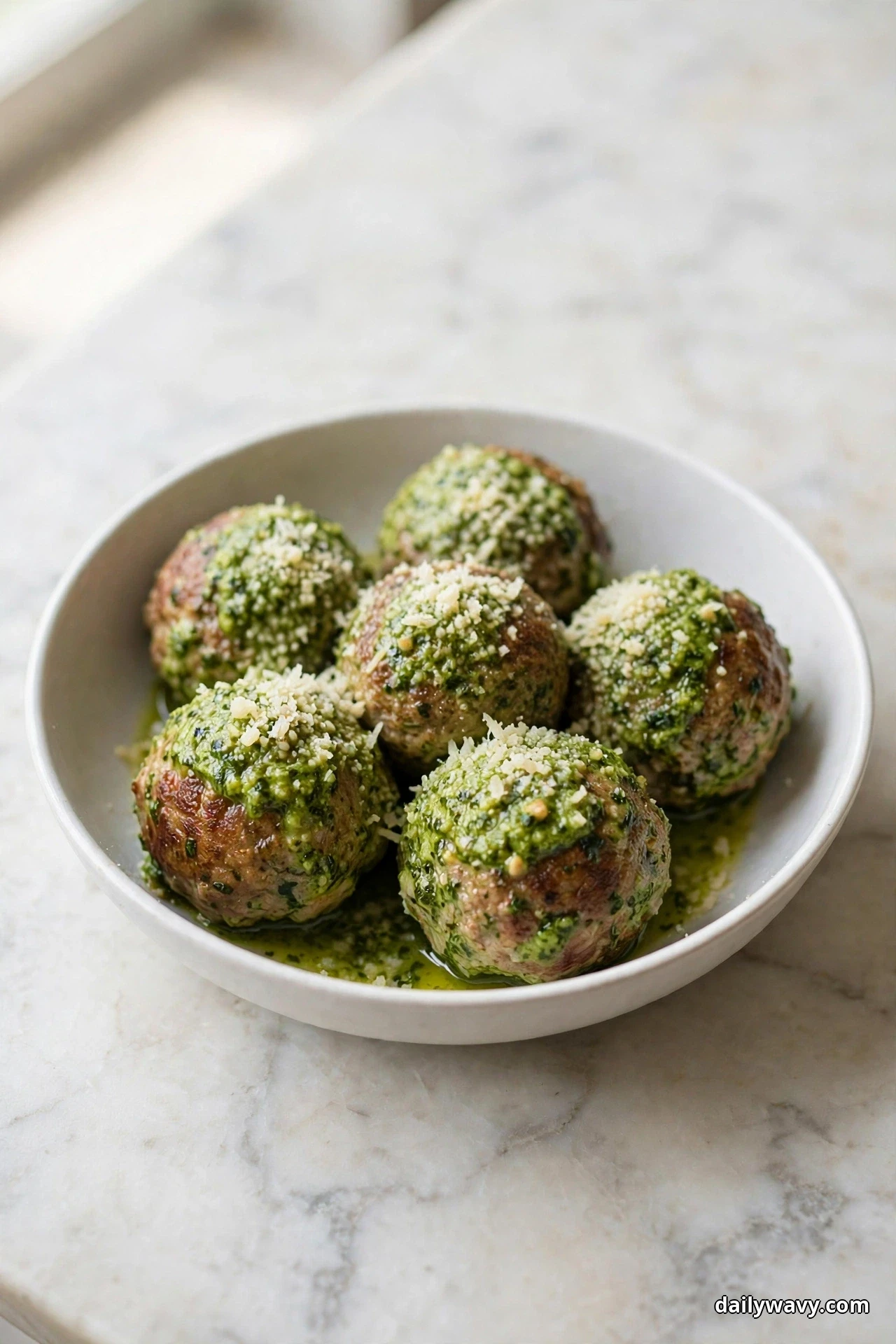 A white bowl holds pork meatballs coated in green basil pesto and dusted with grated Parmesan on a marble surface
