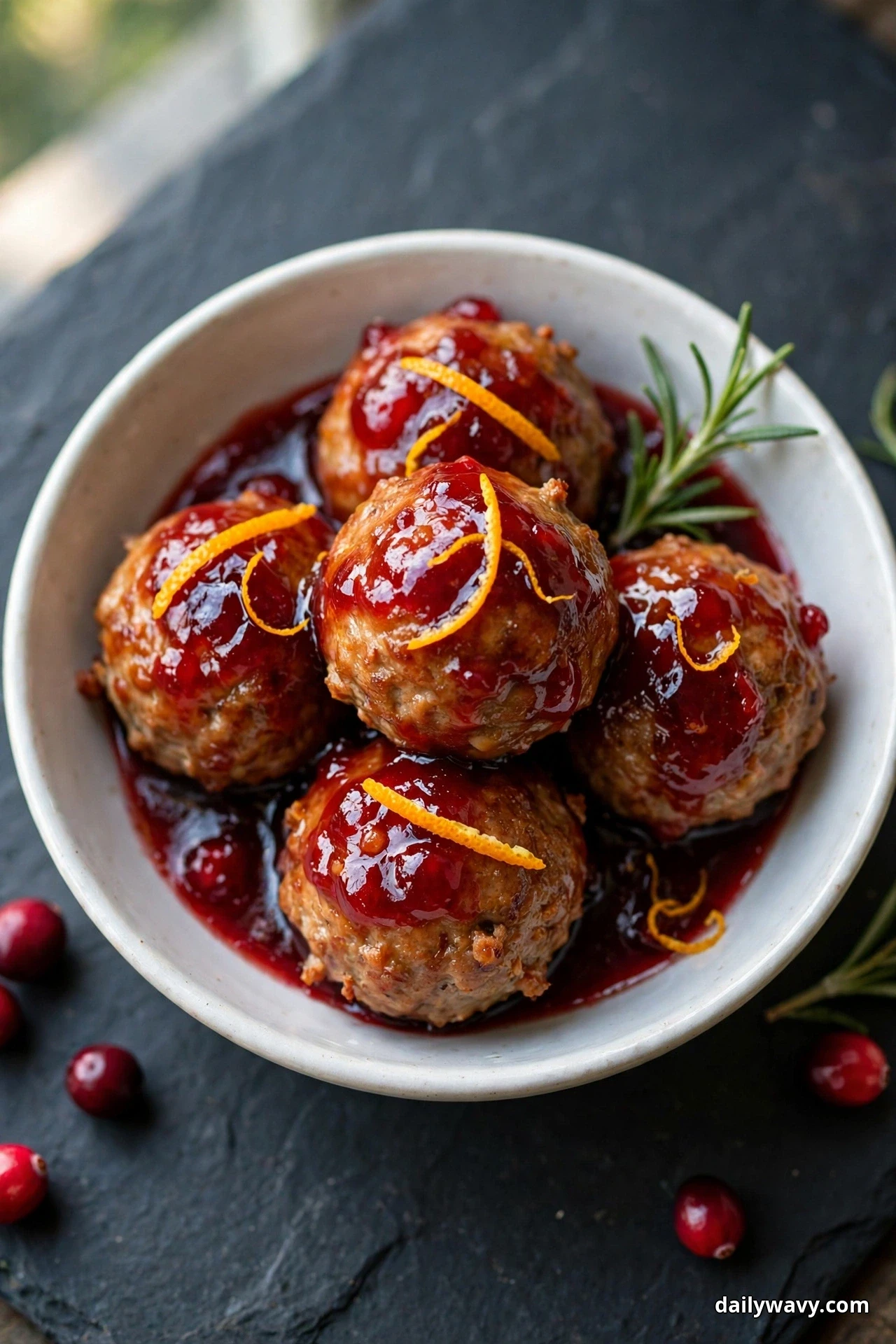Seen from above, a white bowl holds pork meatballs coated in a glossy jewel-red cranberry glaze topped with thin strips of orange zest, on a dark slate board