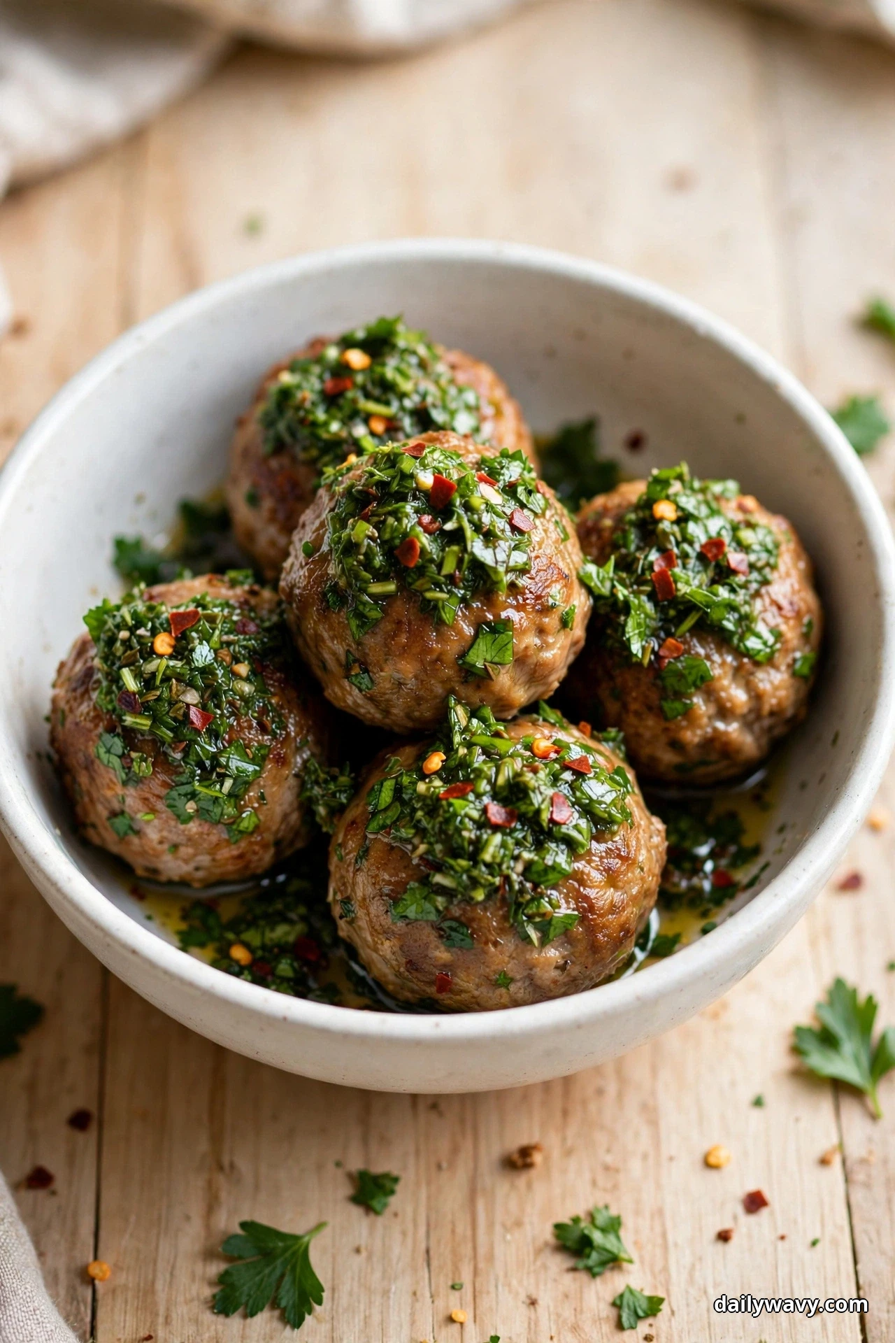 A white bowl holds pork meatballs heaped with chopped green chimichurri flecked with red pepper on a light wooden table