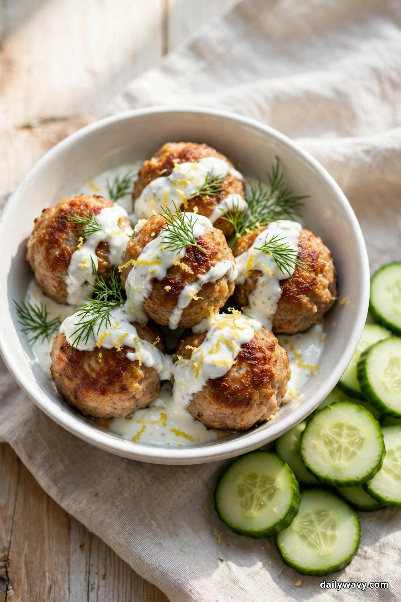 A white bowl holds pork meatballs drizzled with a white lemon herb yogurt sauce and topped with dill fronds and lemon zest, with cucumber slices beside the bowl on a linen cloth
