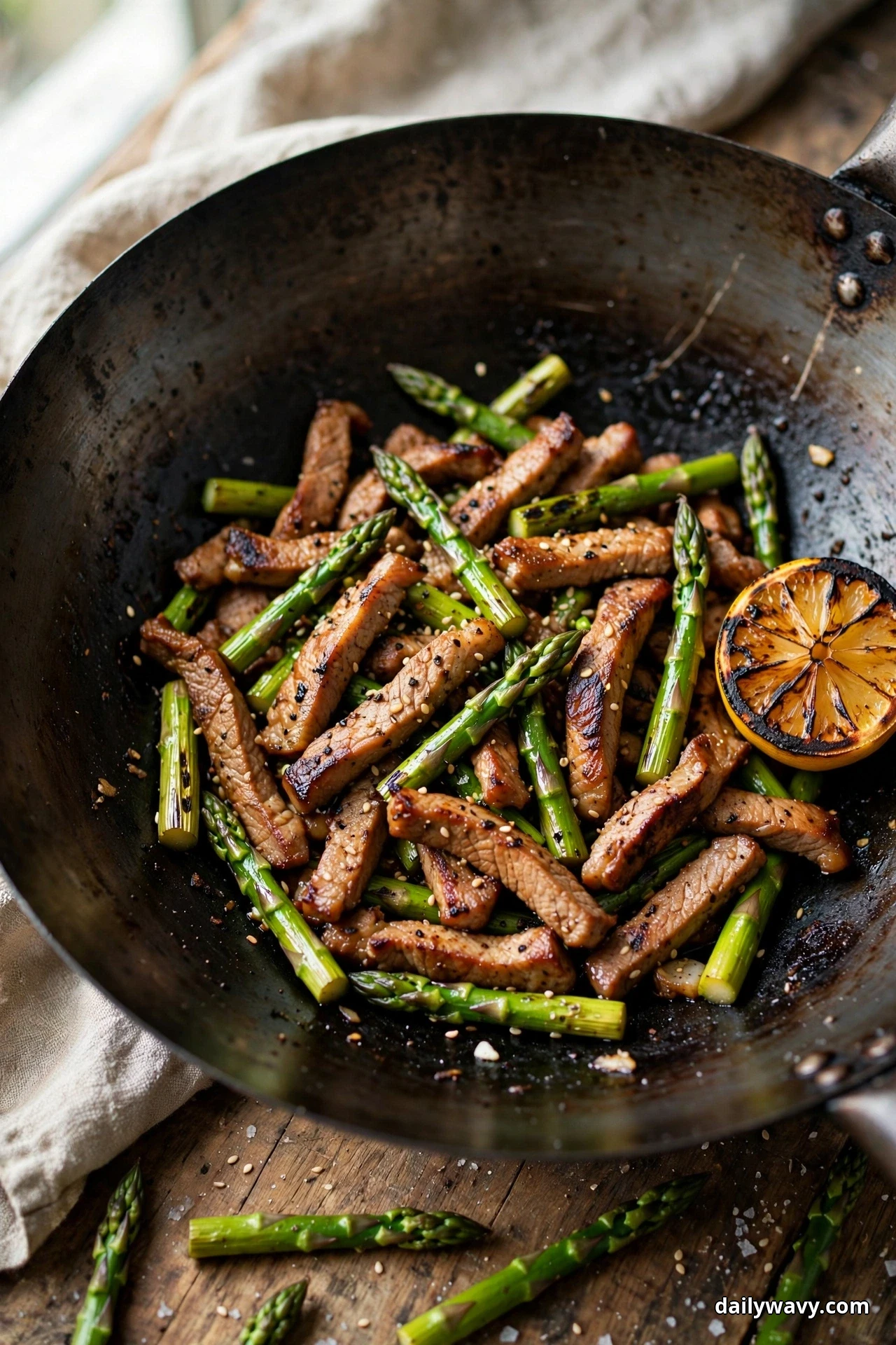 A dark wok holds seared pork strips and asparagus pieces sprinkled with sesame seeds beside a charred lemon half, with raw asparagus spears scattered on the wooden table below