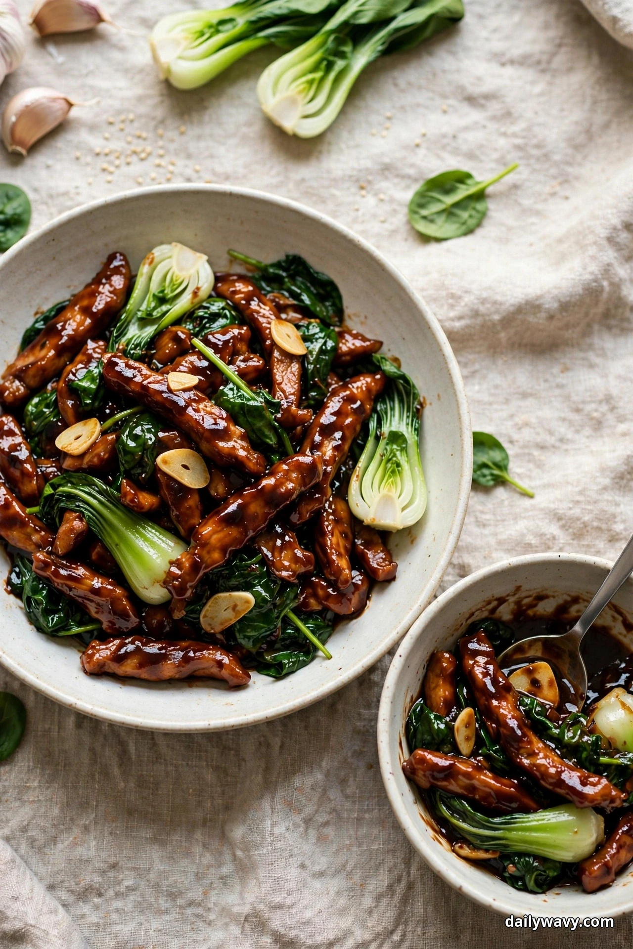 A wide bowl of glossy hoisin-glazed pork strips with wilted spinach, bok choy and garlic slices sits on linen beside a smaller served bowl with a spoon
