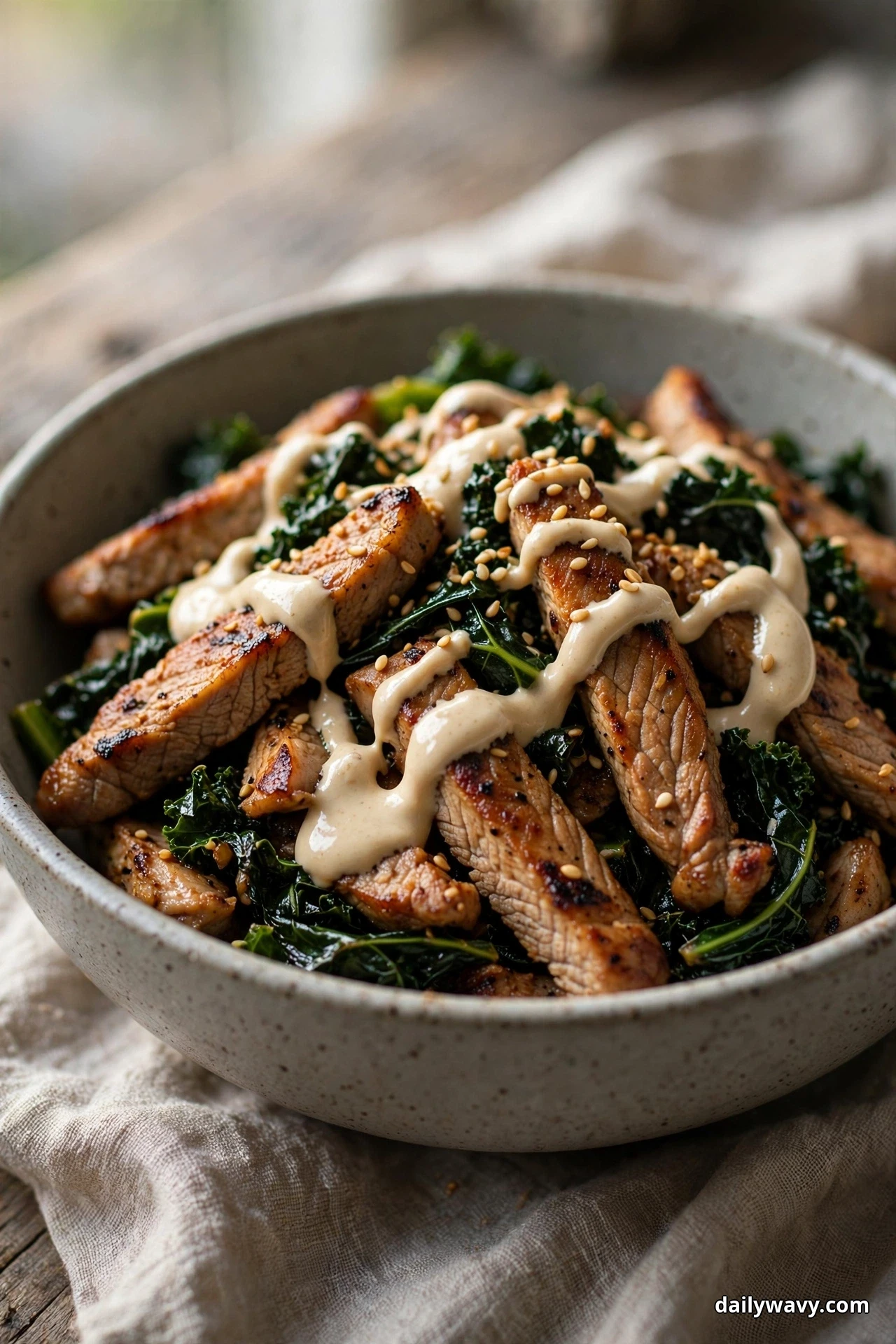 A ceramic bowl of seared pork strips and wilted kale is drizzled with creamy tahini sauce and sprinkled with sesame seeds