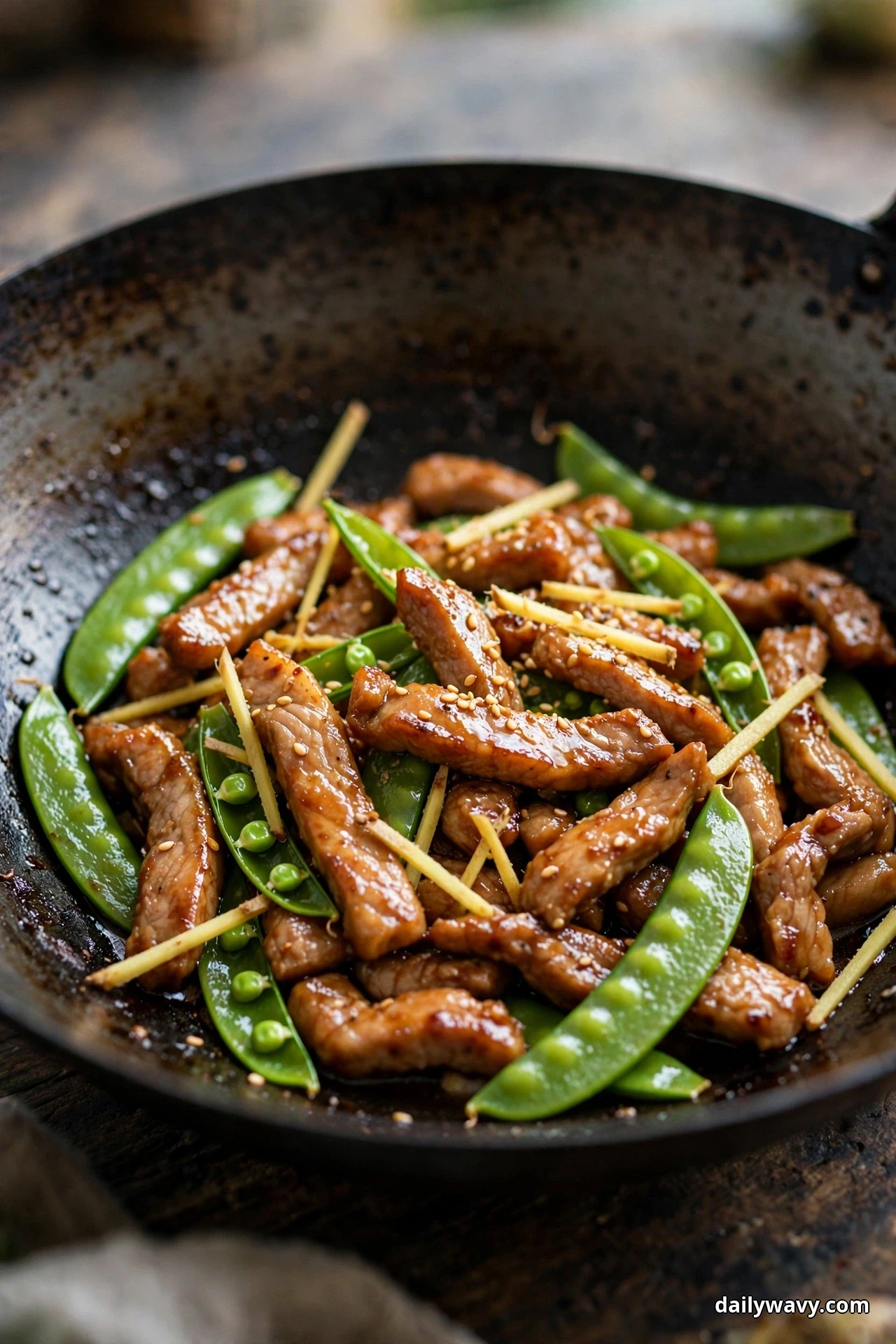 A dark wok holds glossy pork strips, bright sugar snap peas and pale ginger matchsticks on a wooden table