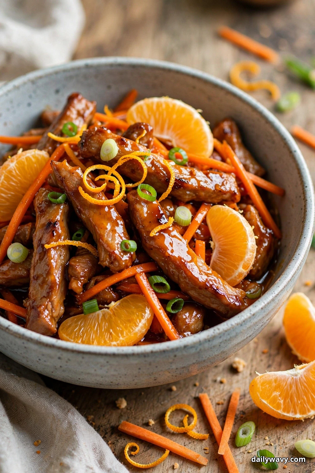 A ceramic bowl of glossy pork strips and julienned carrots is topped with orange wedges, curls of orange zest and scallion rings, with more orange segments on the wooden table beside it